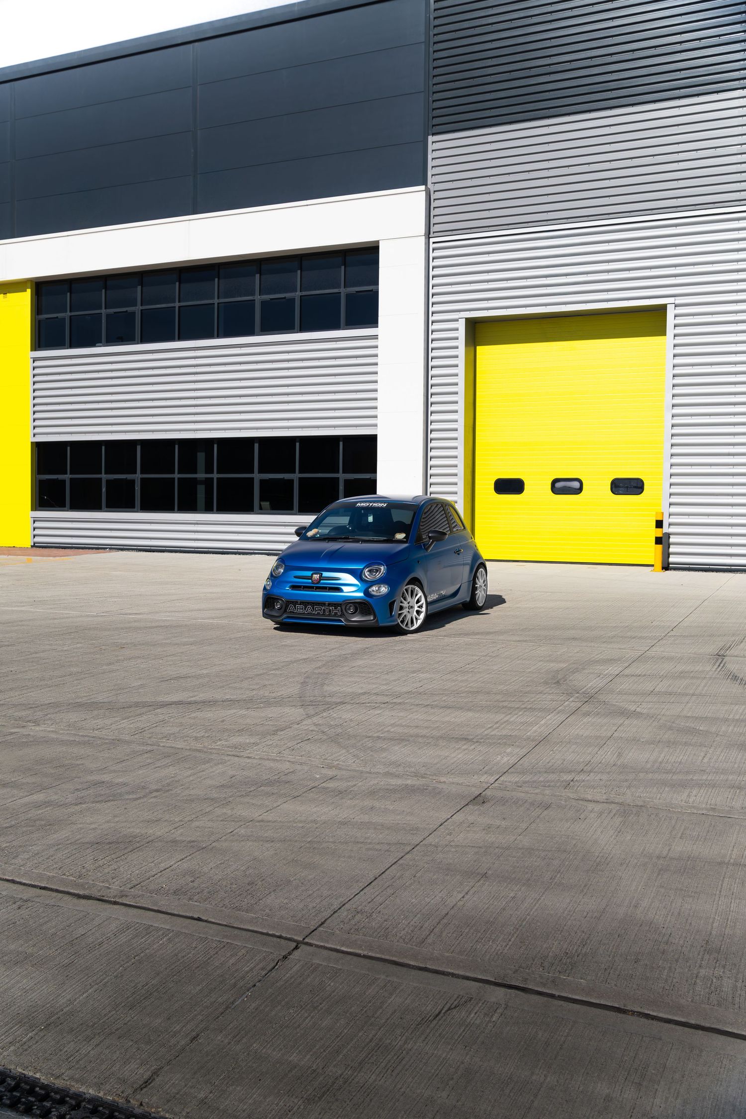 Modified blue Abarth 500 photographed in a wide industrial yard with yellow doors.