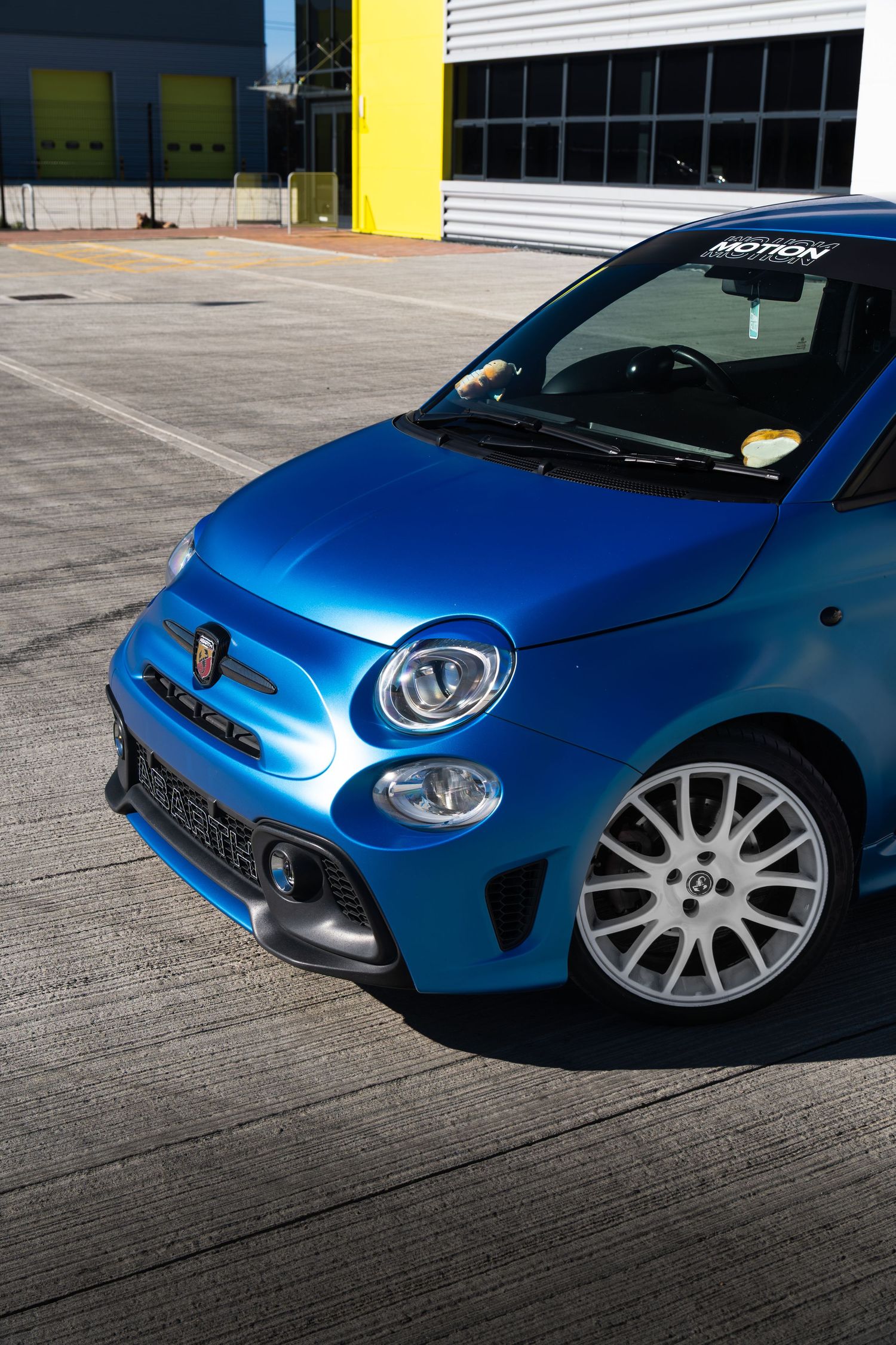 Front close-up of blue Abarth 595 showing alloy wheel, bumper and Motion windscreen sticker.