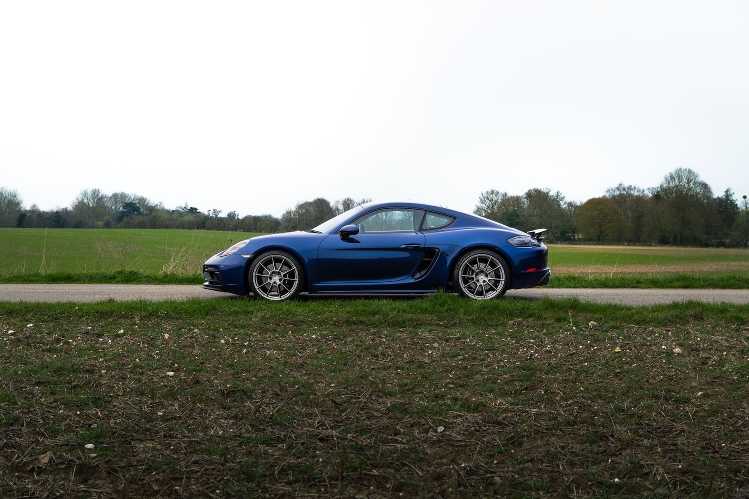 Sapphire blue Porsche 718 Cayman GTS photographed from the side on a countryside road with trees in background.