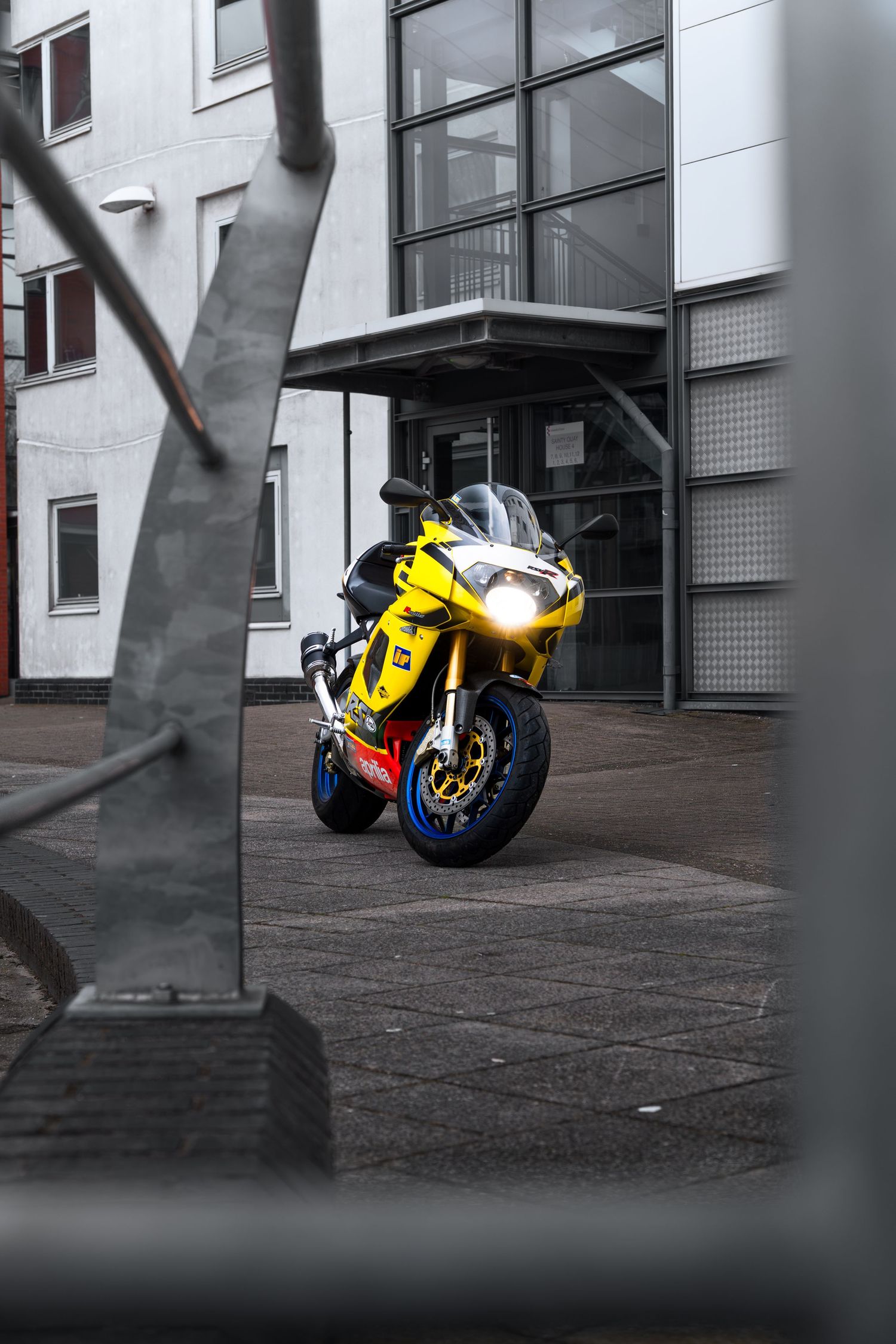 Yellow Aprilia RSV1000 sportbike photographed through metal railings outside a gray building entrance.