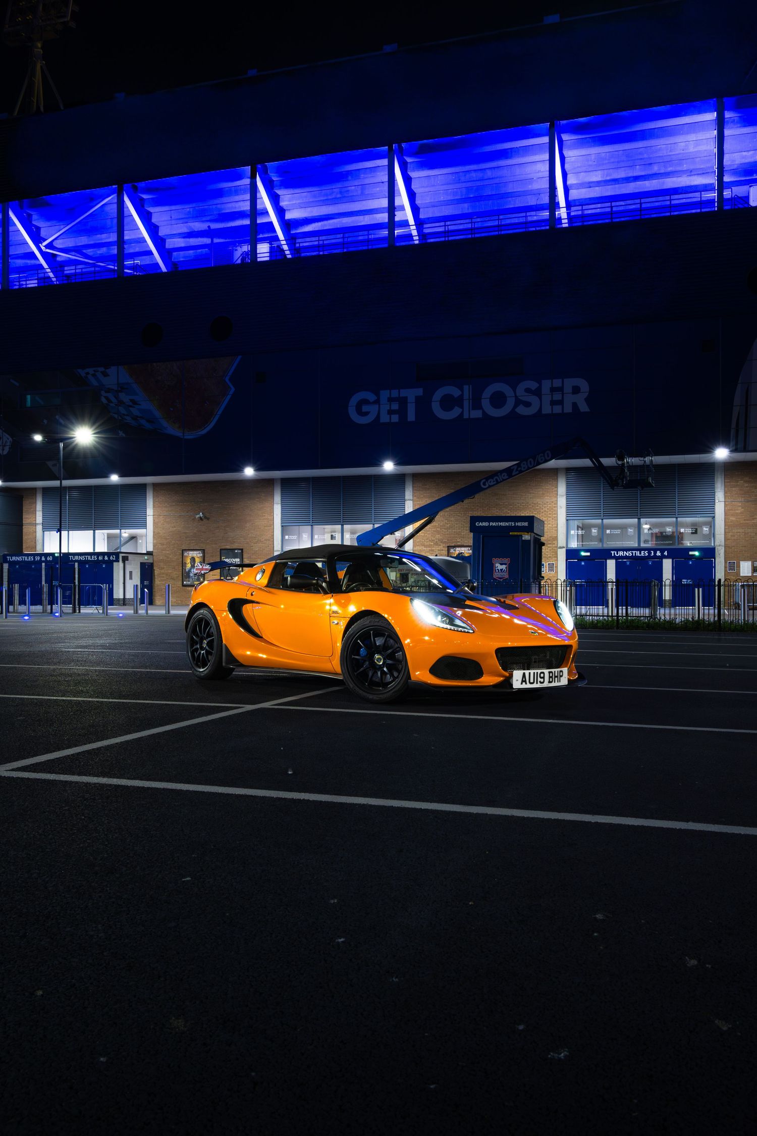 Orange Lotus Elise sports car with open door in dark stadium car park under blue-lit GET CLOSER signage.