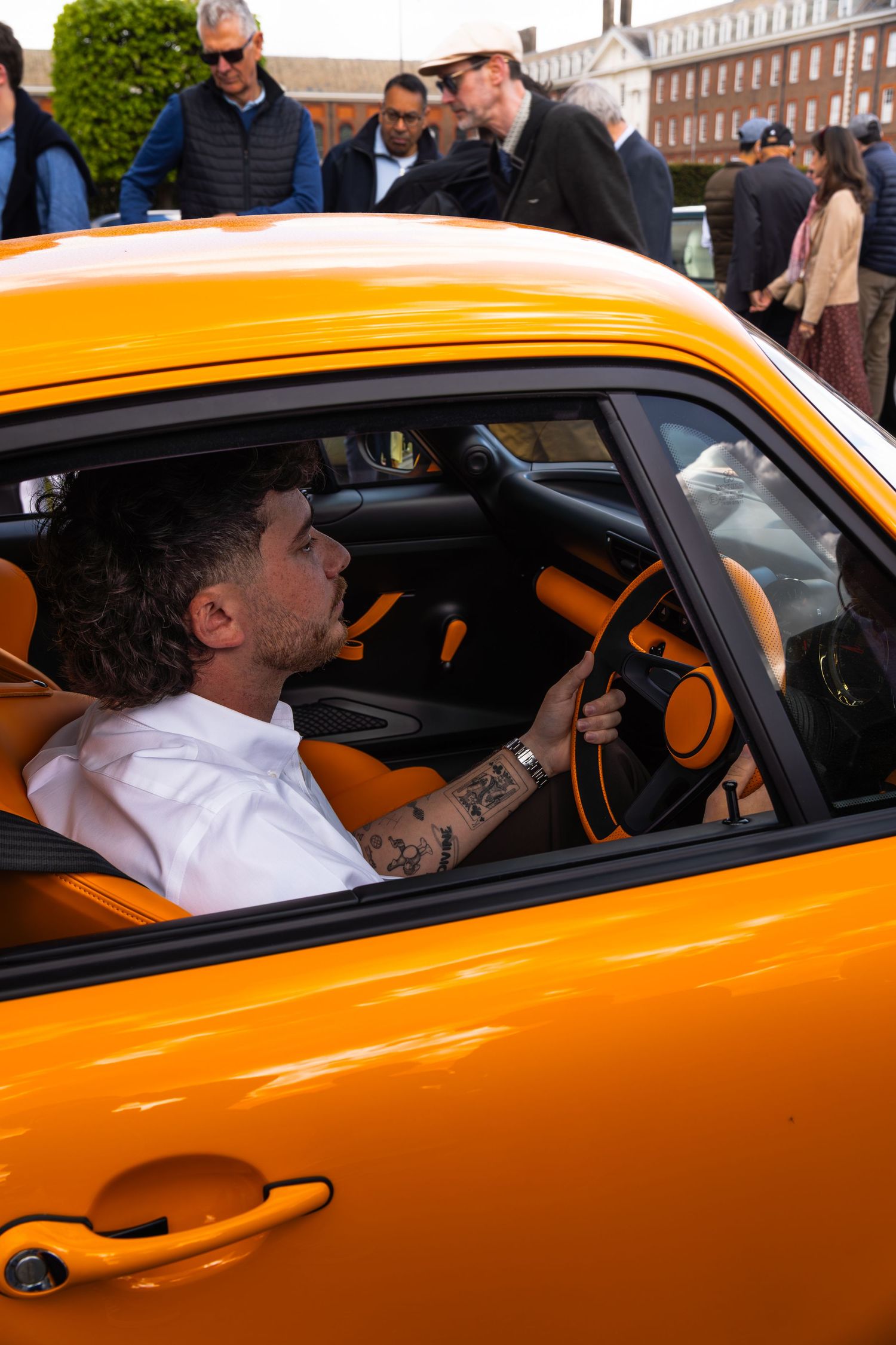 Tattooed man driving orange Porsche with matching orange interior at an outdoor car event.