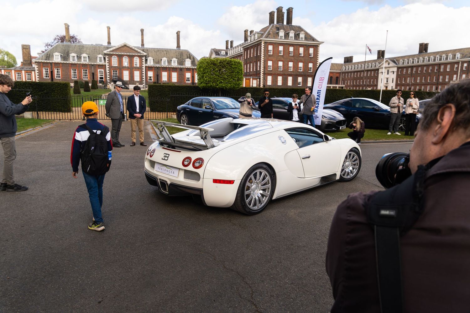 Rear view of a white Bugatti Veyron supercar at an outdoor car show with a historic building backdrop.