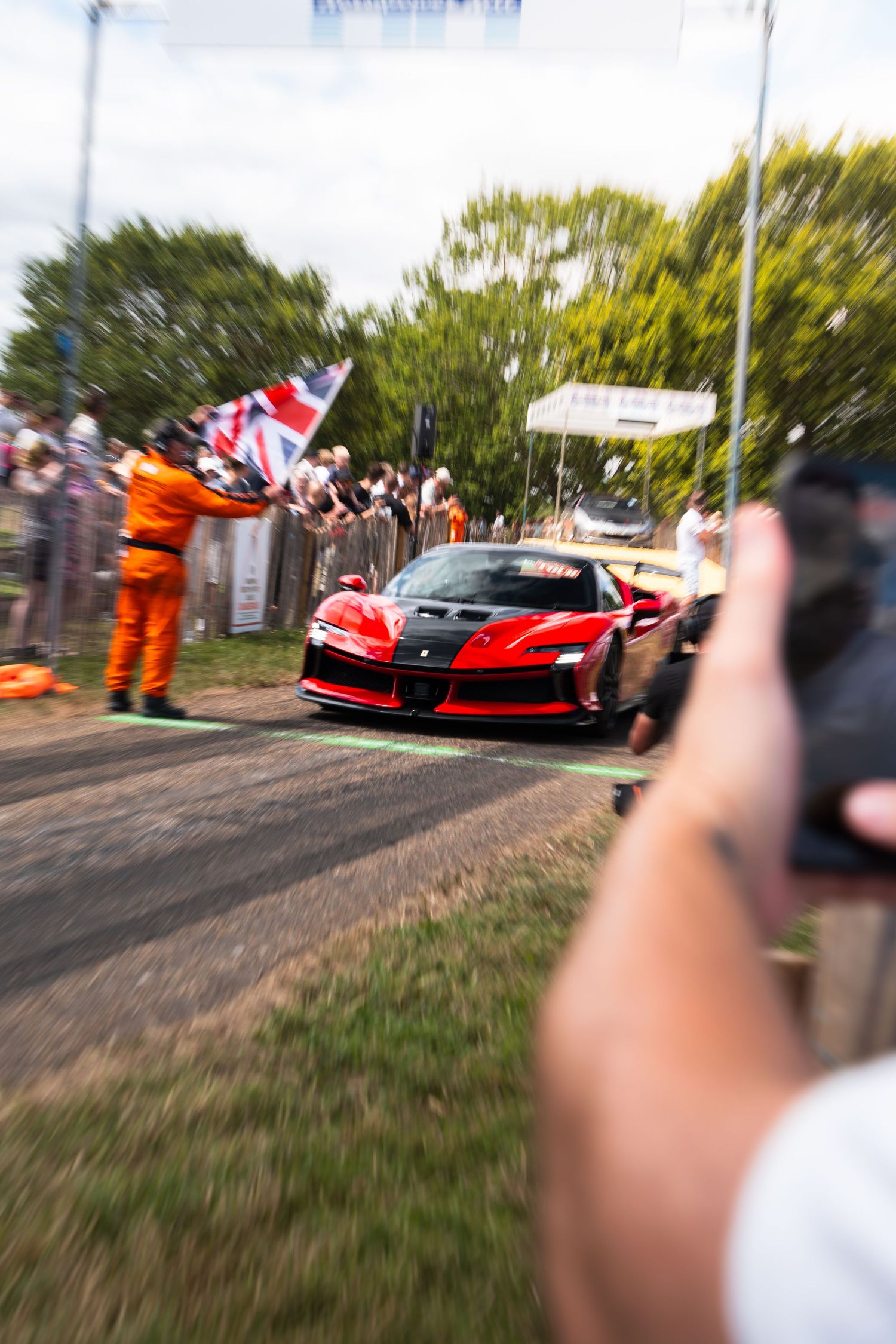 Red Ferrari supercar at Goodwood Festival of Speed hillclimb, crowds cheering with flags, motion blur effect.