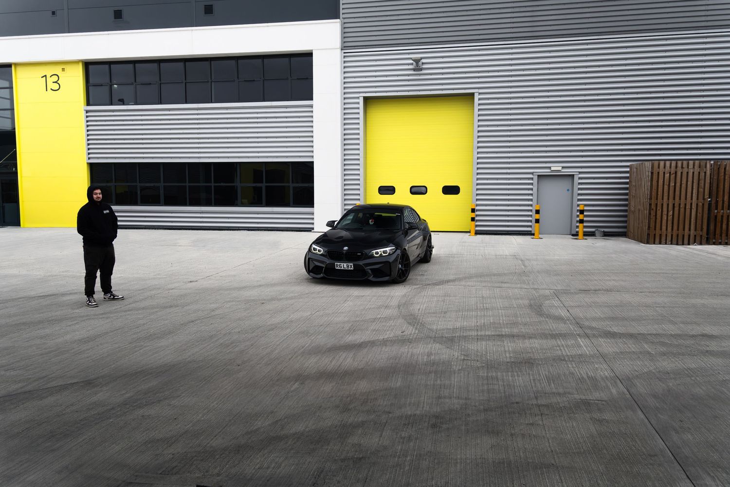 Black BMW M2 performing donuts in a large industrial yard with yellow garage doors.