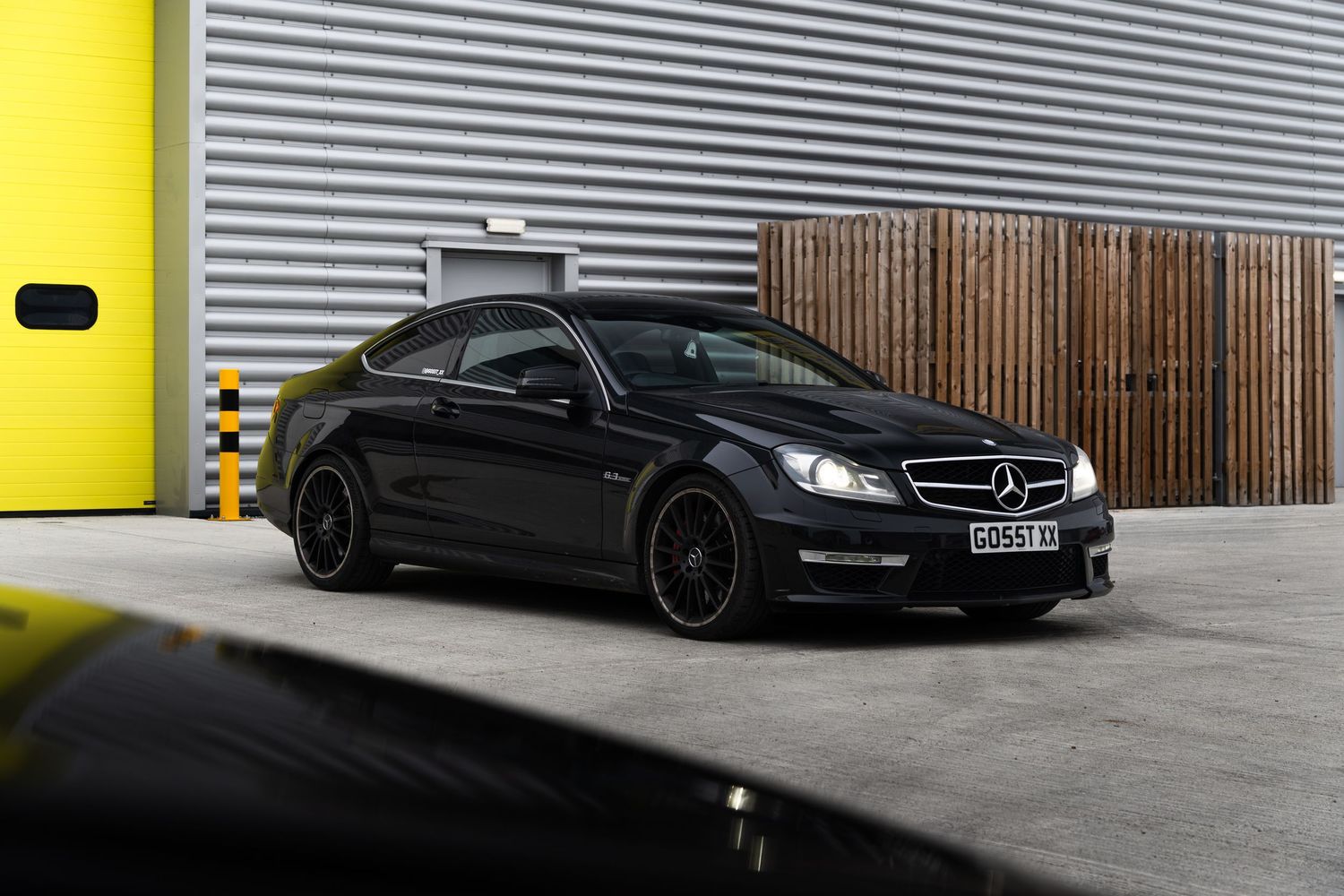Black Mercedes-Benz C63 AMG coupe with dark alloy wheels parked outside grey corrugated metal industrial building.