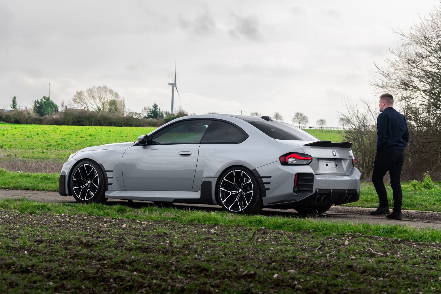 Silver BMW M2 coupe with wide body kit parked on a rural path as a man admires it from behind.