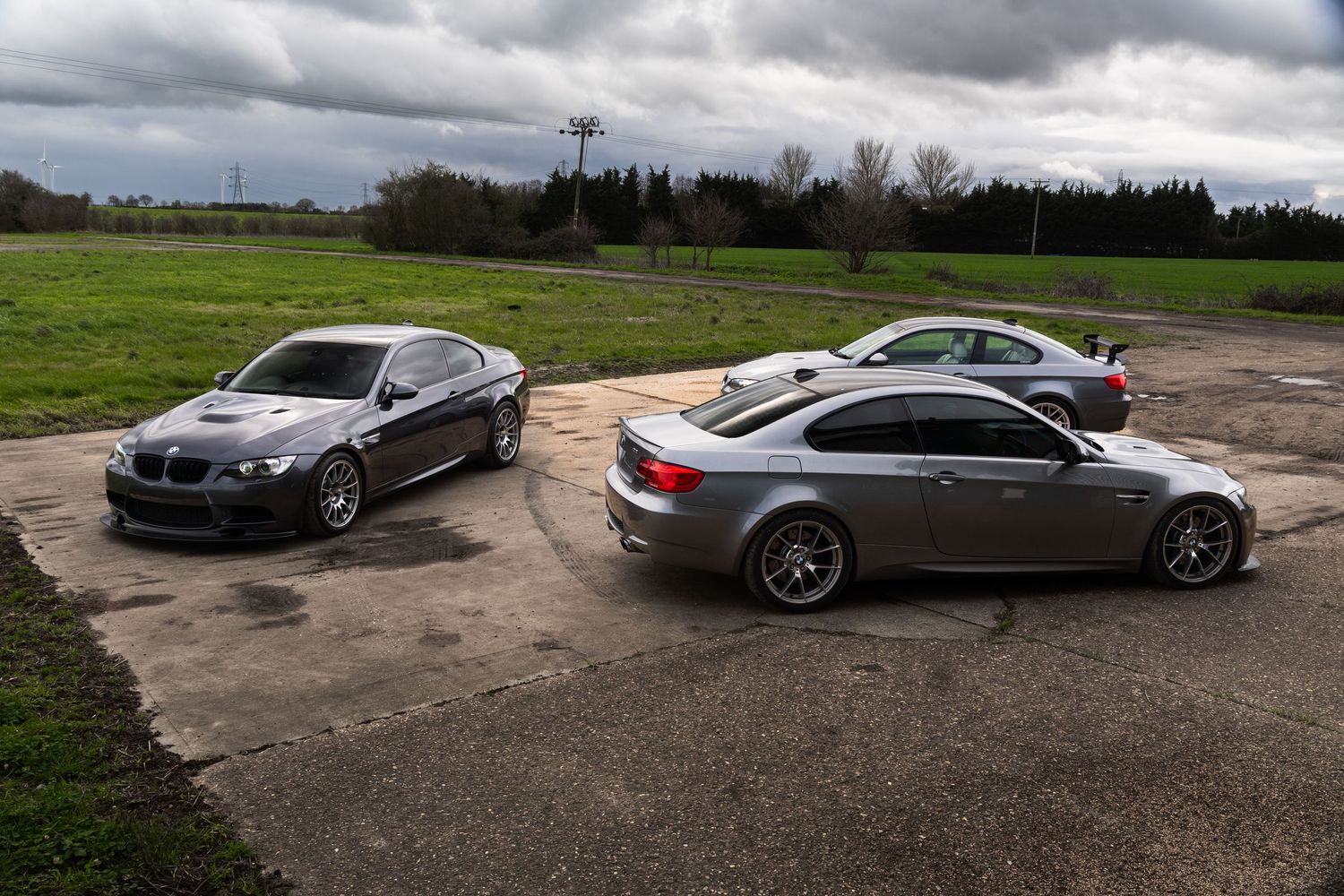 Three gray BMW M3 E92 coupes parked together on a gravel lot with green fields in the background.