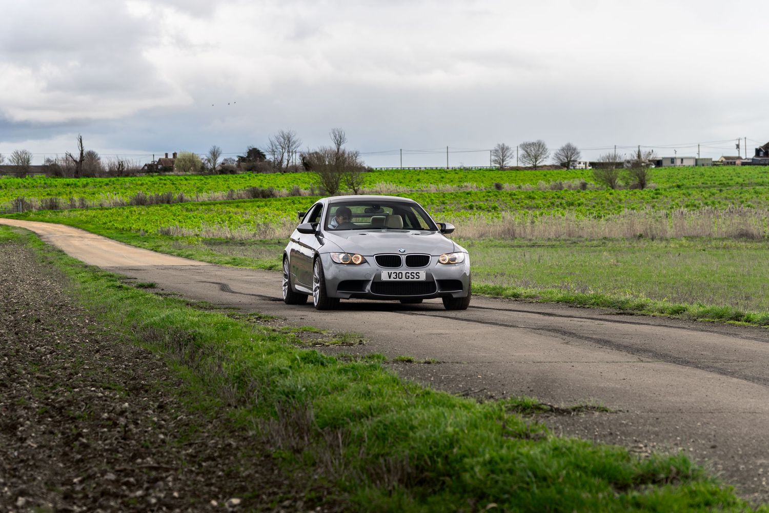 Silver BMW M3 sedan driving along a narrow rural road surrounded by green fields.