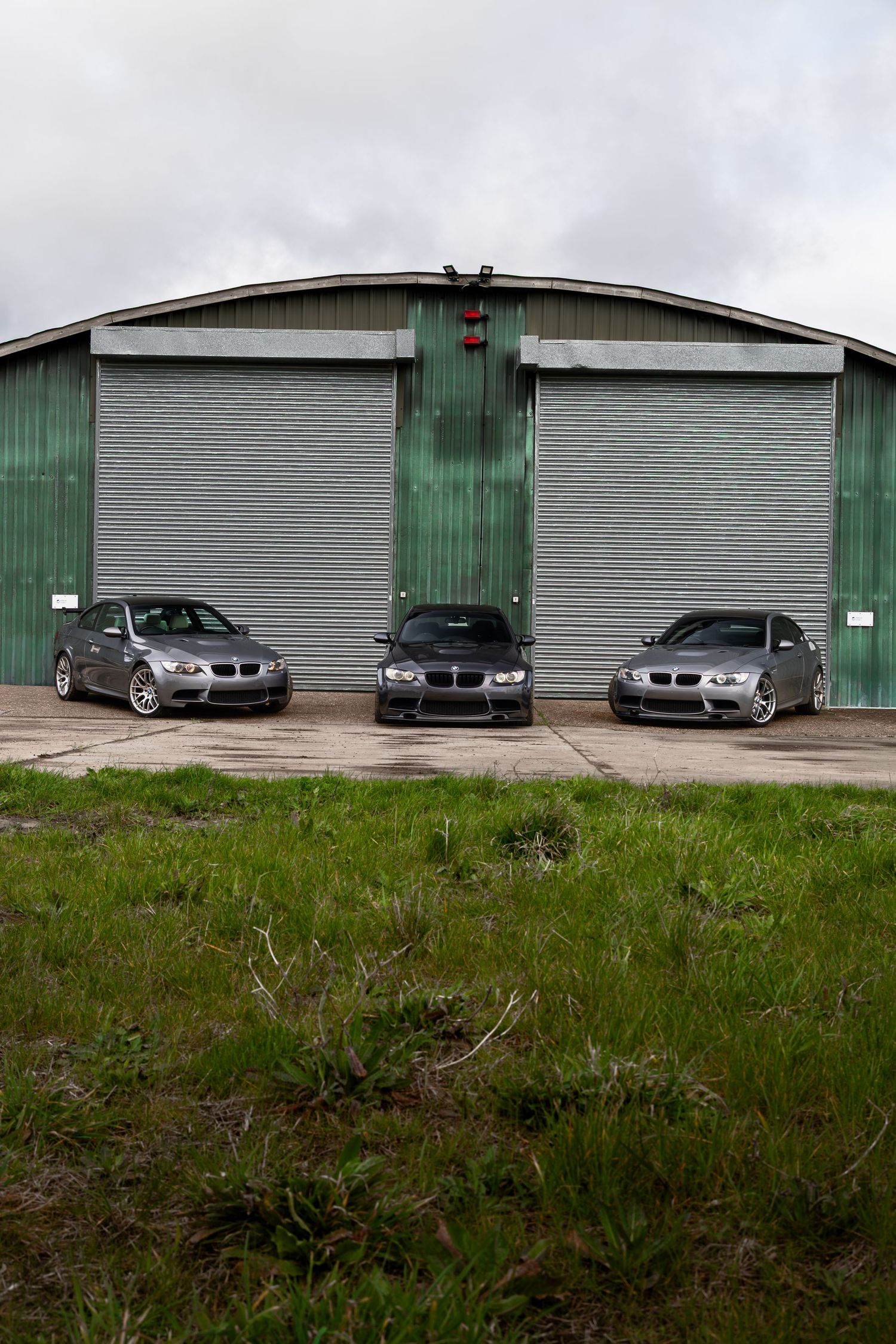 Three grey BMW M3 vehicles parked outside a large green metal warehouse under cloudy skies.