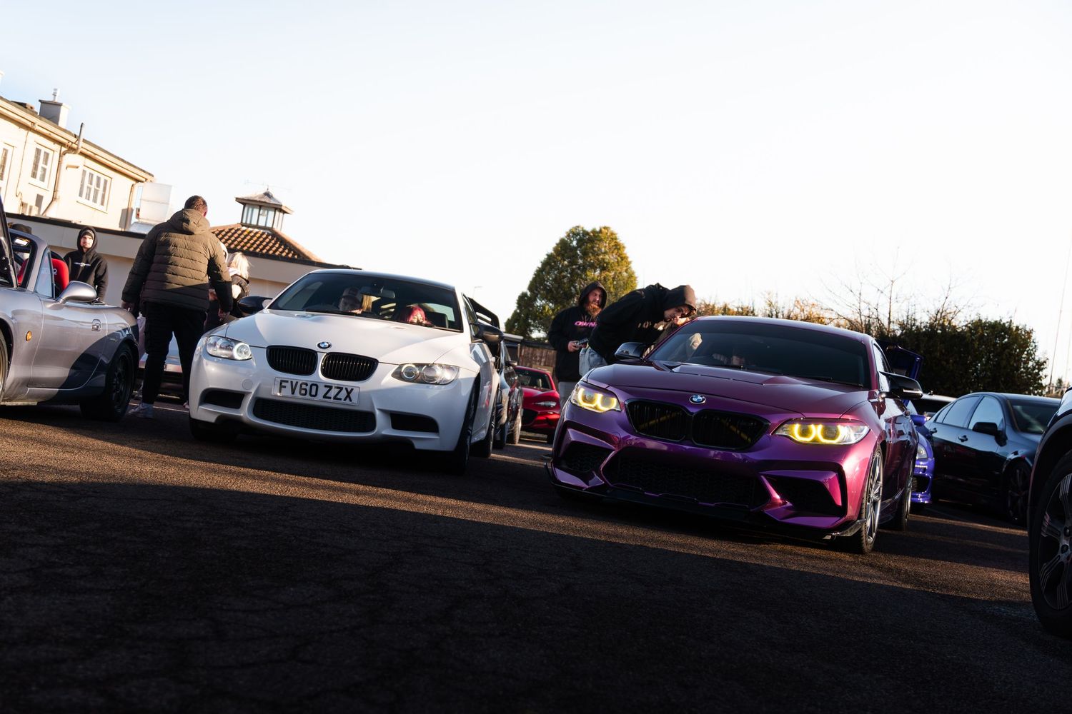 White BMW M3 and purple BMW M2 parked side by side at an outdoor car meet at sunrise.