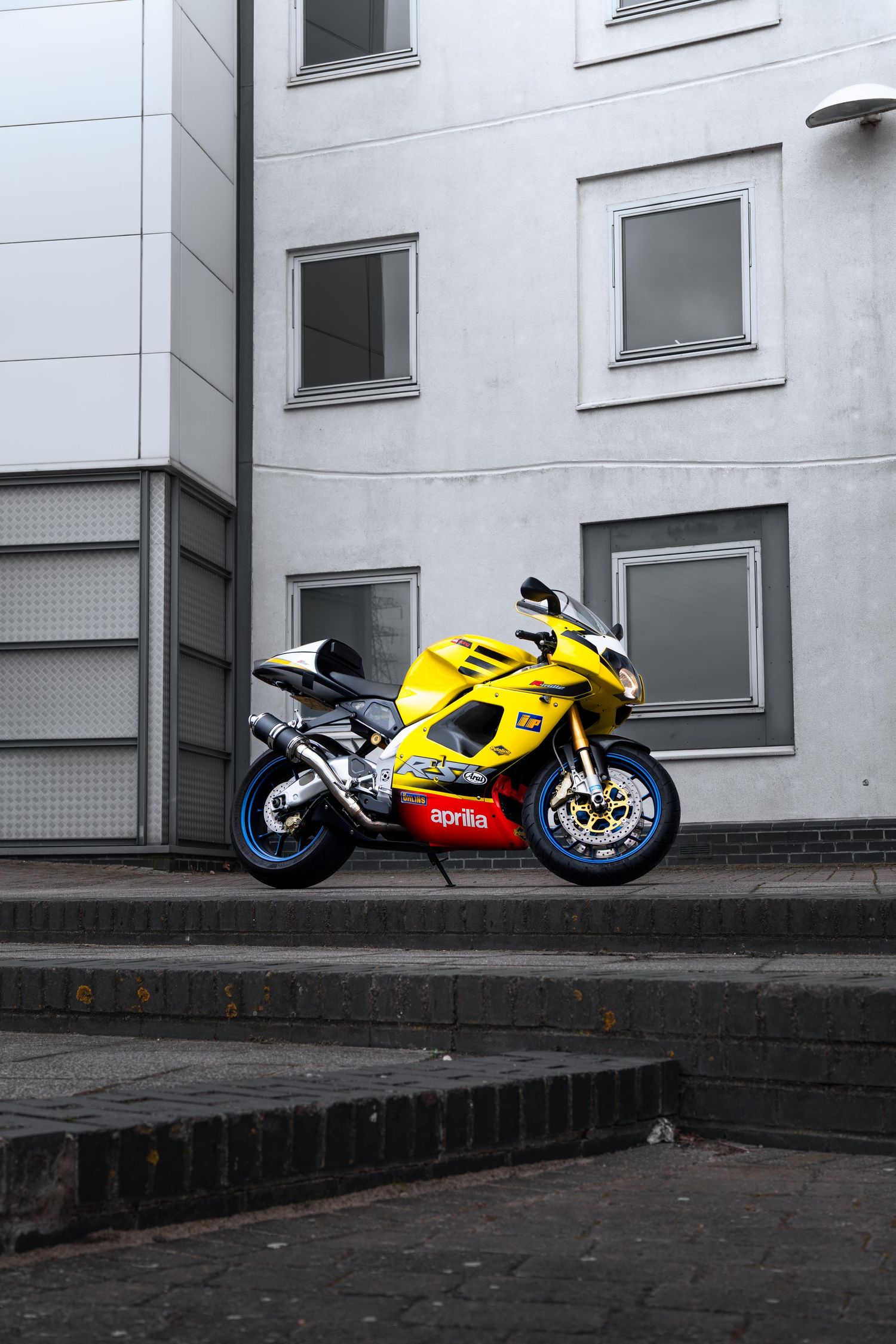 Yellow Aprilia RSV1000 superbike parked on urban steps beside a gray and white building facade.
