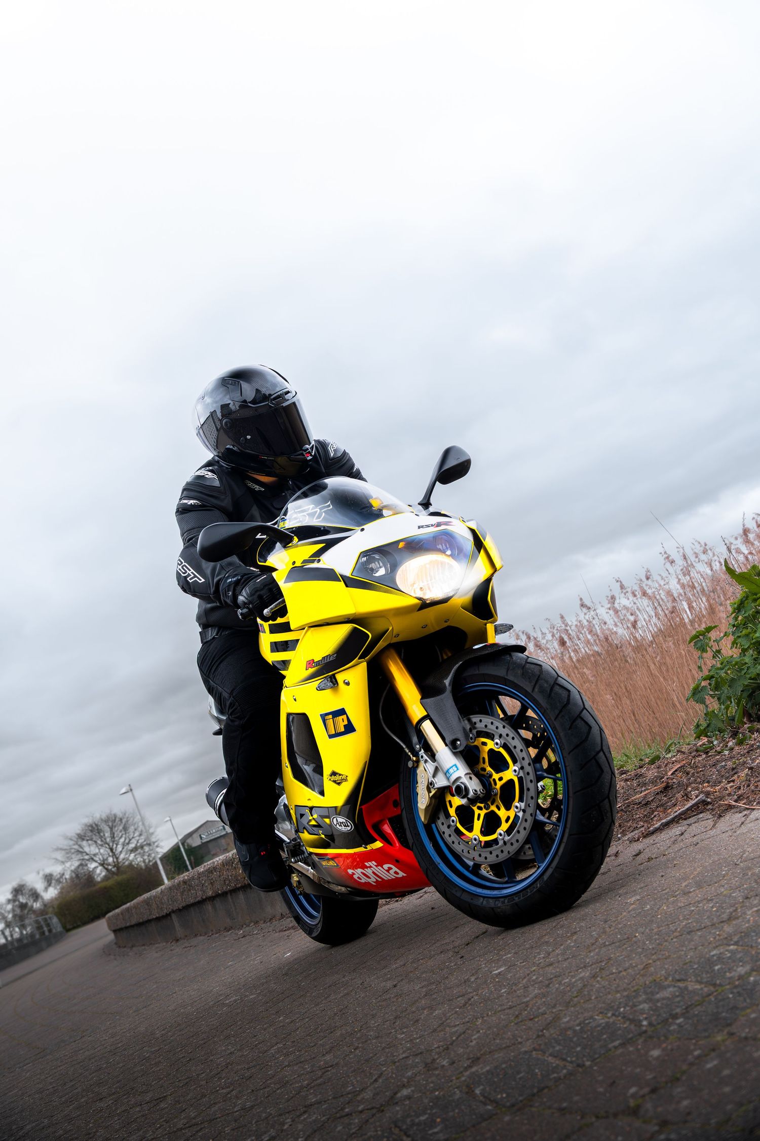Wide-angle front view of a yellow Aprilia sportbike with rider in black gear on a road.