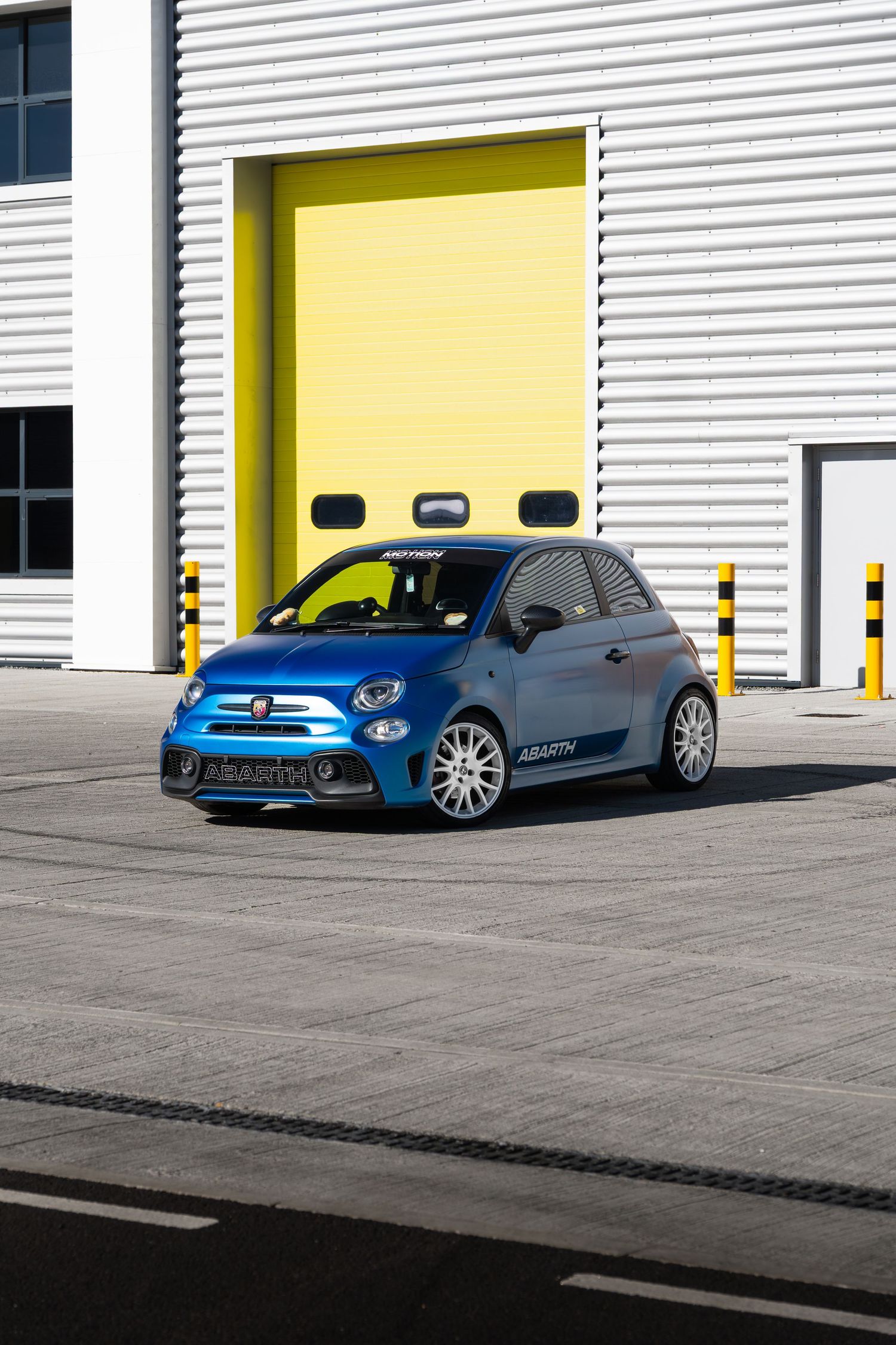 Blue Abarth 595 with Abarth side graphics parked outside yellow-doored industrial building.