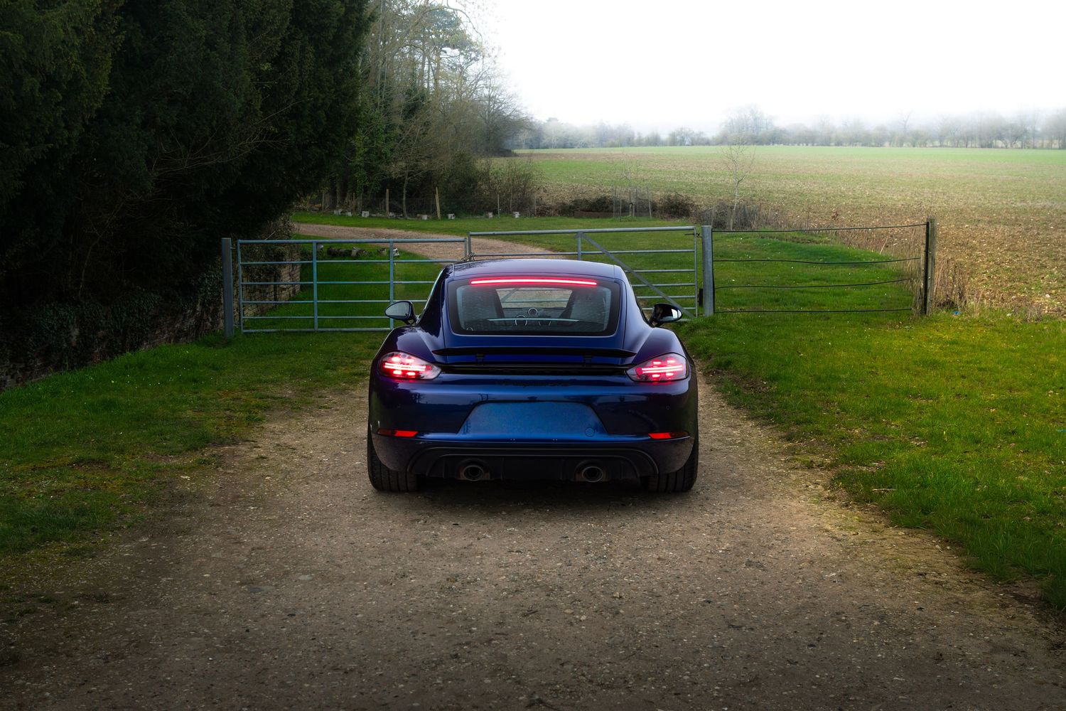 Rear view of a blue Porsche 718 Cayman parked on a gravel path beside a metal farm gate.