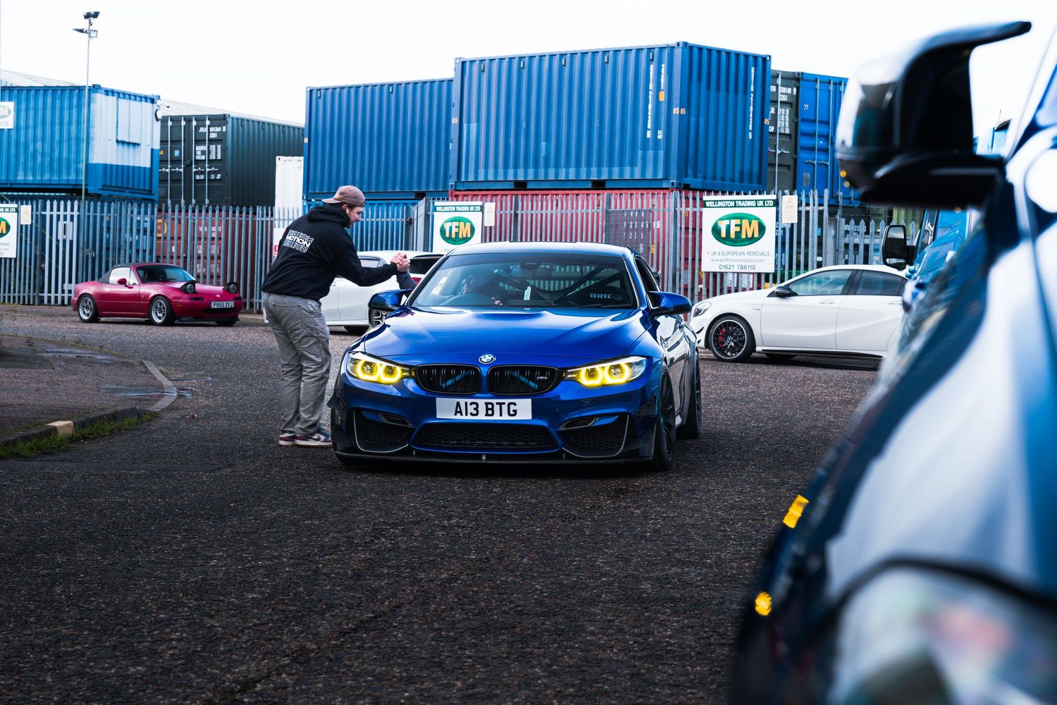 Man admiring a blue BMW M4 with yellow headlights at an outdoor car meet.