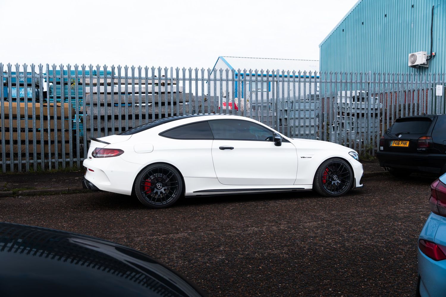 A white Mercedes-AMG C63 Coupe parked near a blue metal fence at a car meet.