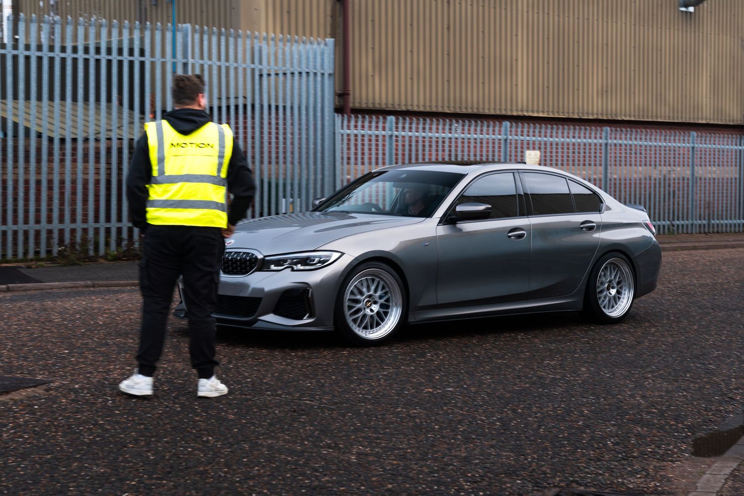 A man in a high-vis vest stands beside a silver BMW 3 Series on a quiet industrial lot.