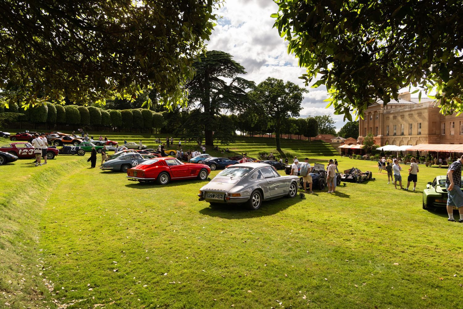 Classic cars displayed on green lawn at elegant country house concours event on sunny day.