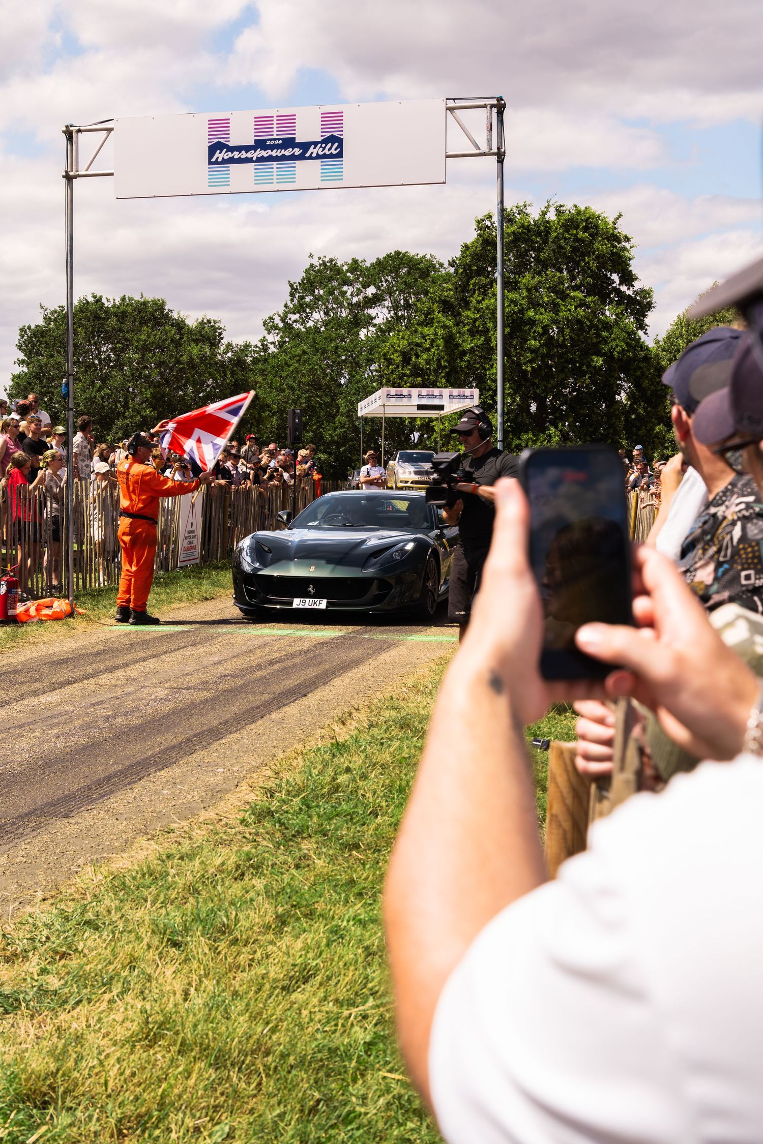 Dark sports car at Goodwood Festival of Speed hillclimb, crowds lining the track, marshal waving flag under banner.