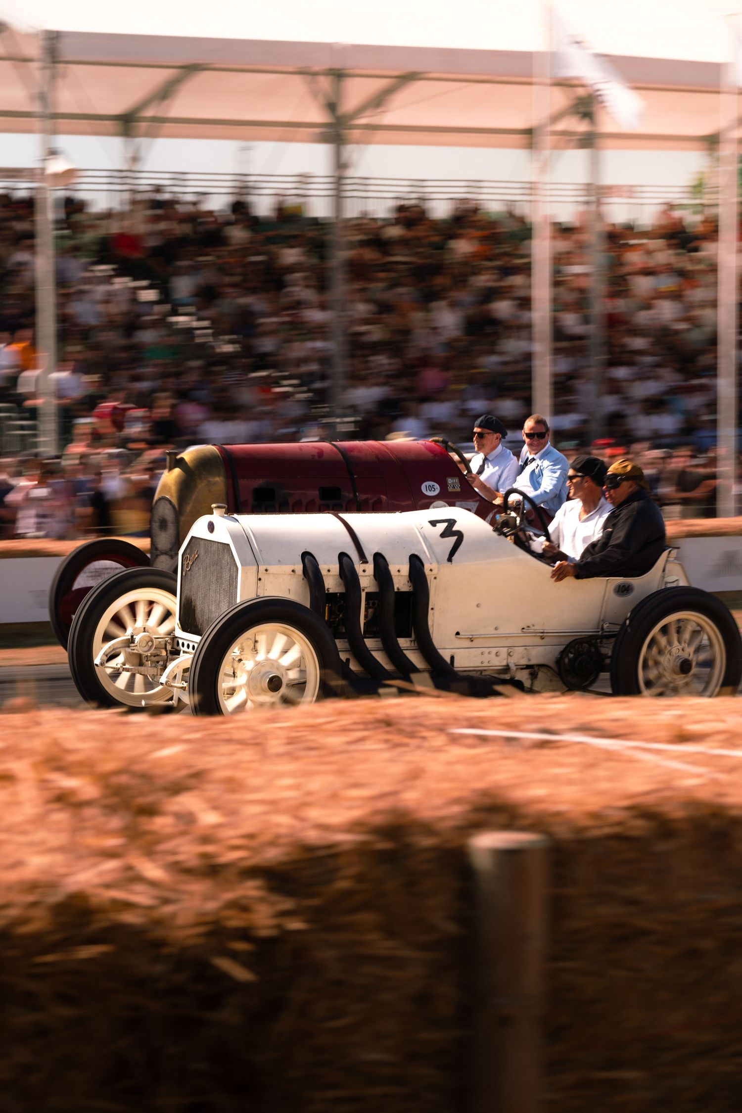 Vintage white race car number 7 with passengers competing at a classic motorsport event with grandstands full of spectators.