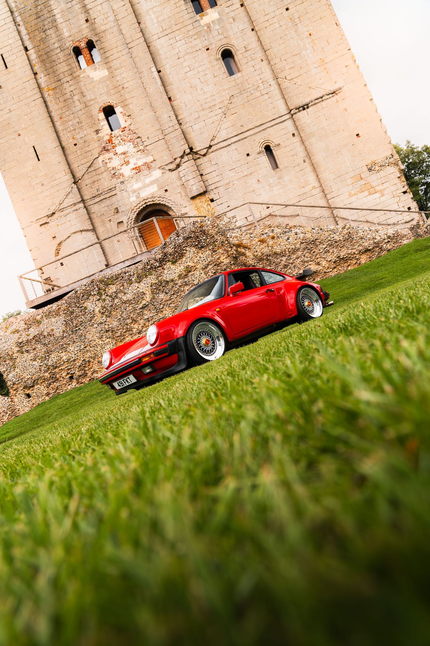 Red Porsche 911 Turbo parked on lush green grass beside a historic stone castle tower on a cloudy day.