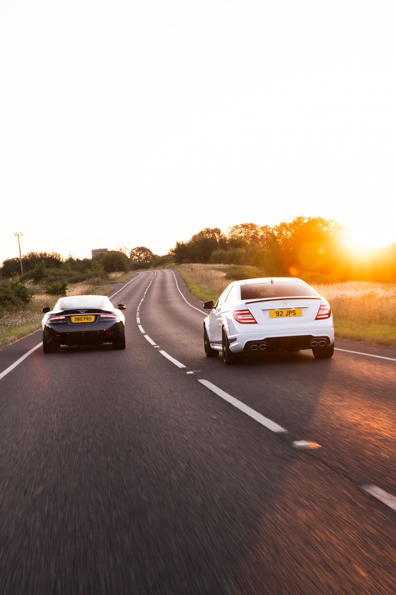 Two sports cars driving on a winding road at golden sunset, white Mercedes leading a dark coupe on a scenic route.