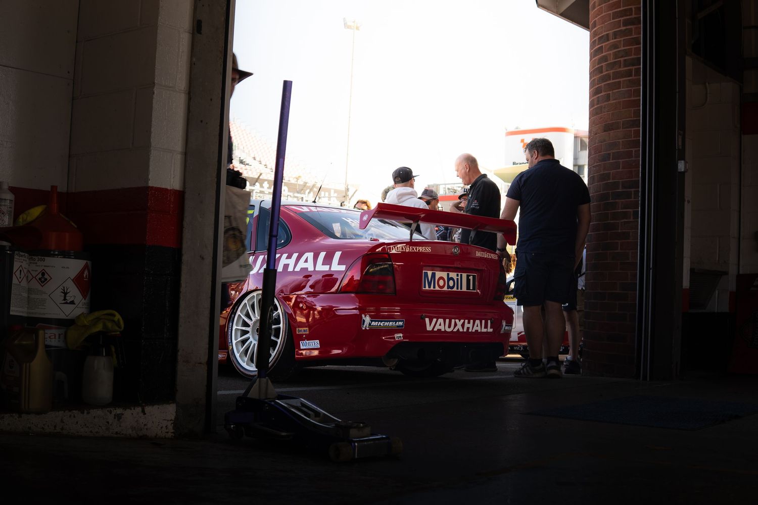 A red Vauxhall race car being worked on by mechanics inside a dimly lit pit garage.