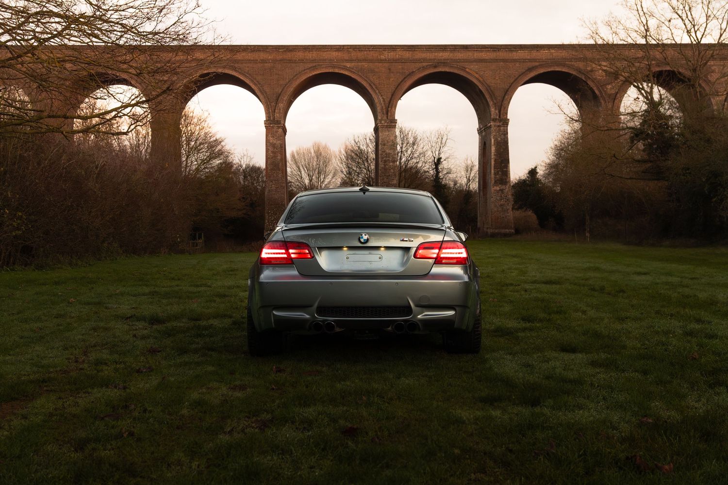 Rear view of a silver BMW M3 parked on grass before a historic brick viaduct at dusk.