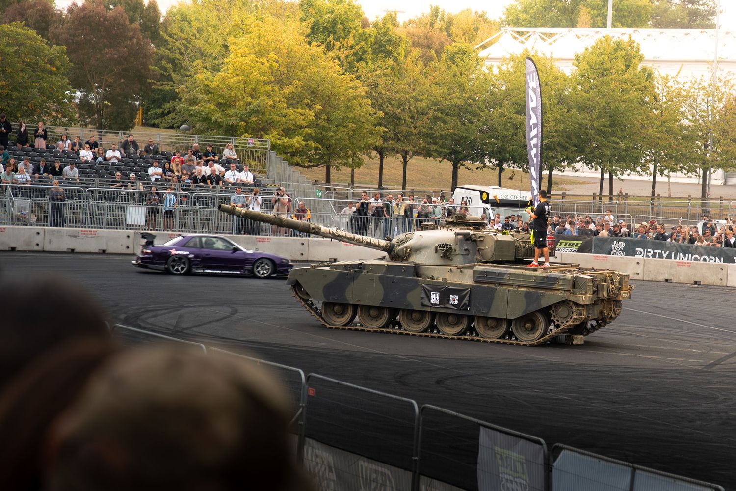 A military tank races alongside a purple sports car on a track in front of a large crowd at an outdoor motorsport event.