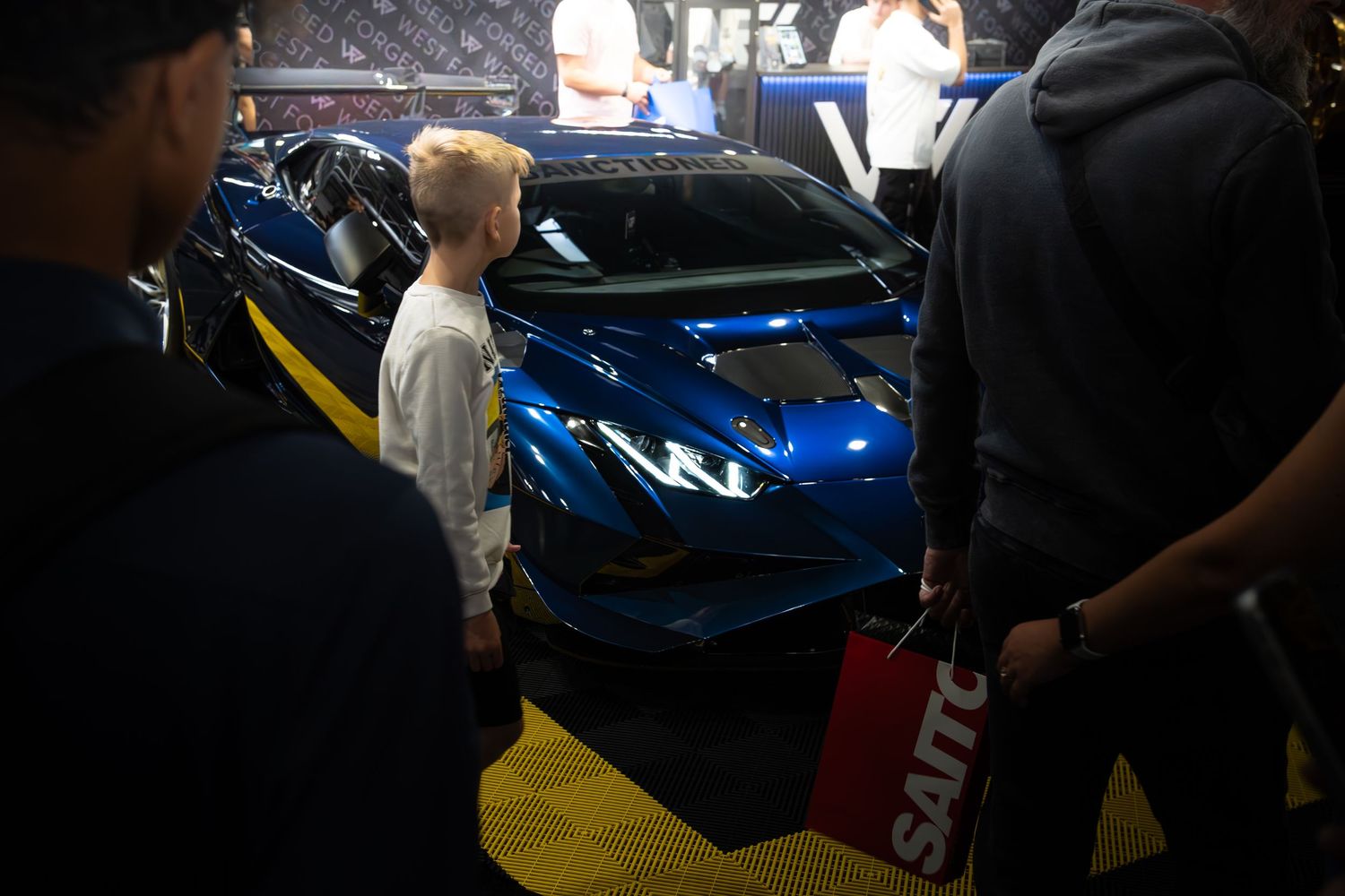 A young boy admires a shiny blue Lamborghini Huracan at an indoor car show.