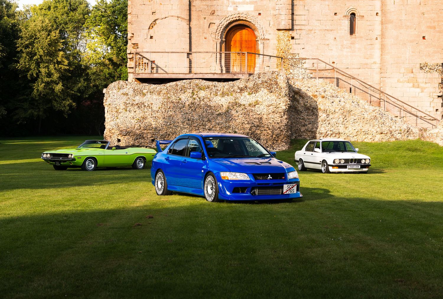 Three classic cars, including a blue Mitsubishi Evo, green muscle car, and white BMW, parked on grass before a stone building.