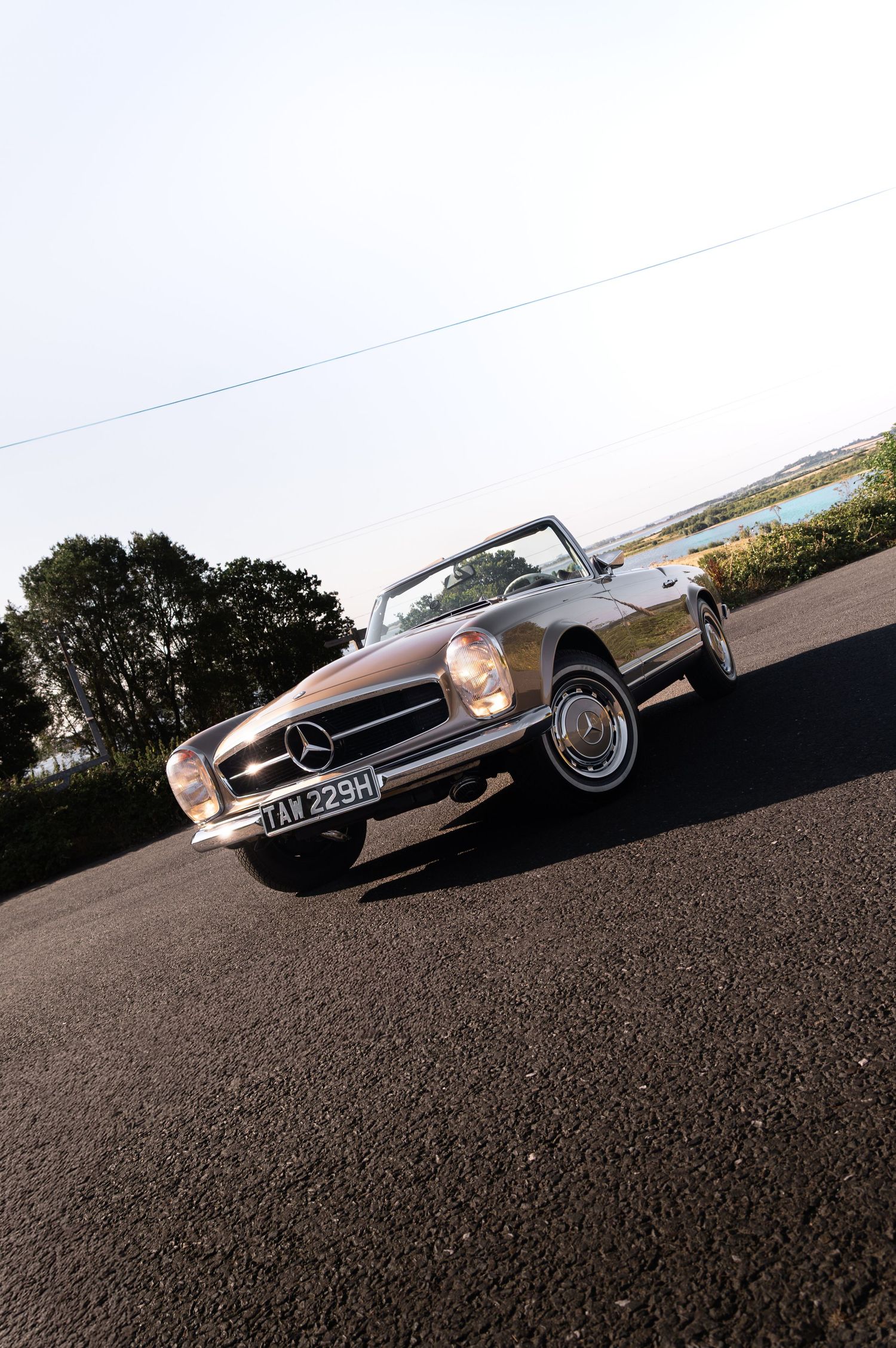 Classic Mercedes-Benz convertible sports car in gold, parked on tarmac with trees and water in background.