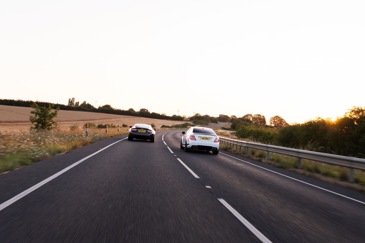 Two cars driving on a dual carriageway at sunset with golden fields and trees lining the road.