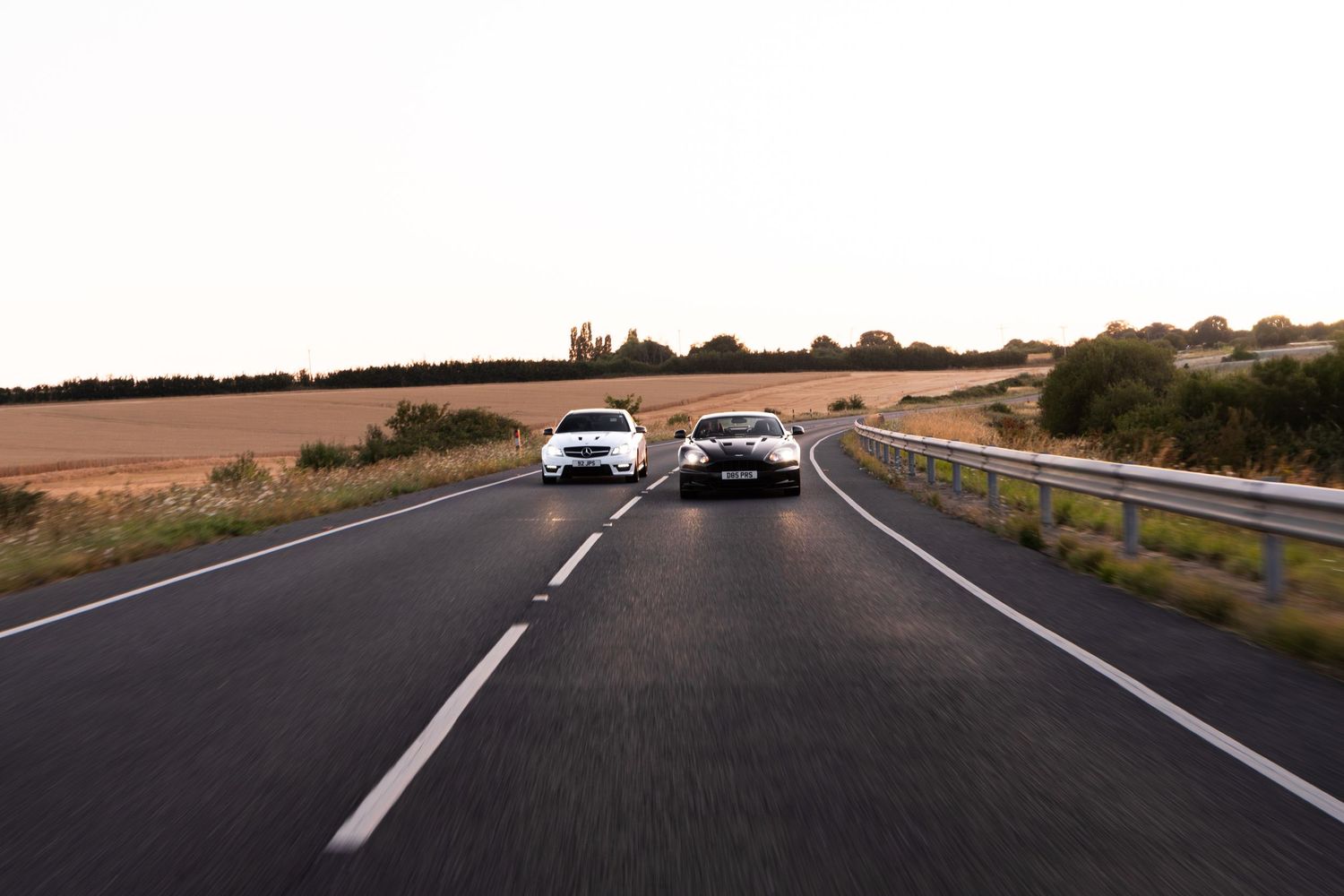 Two cars driving side by side on a rural highway surrounded by fields at dusk.
