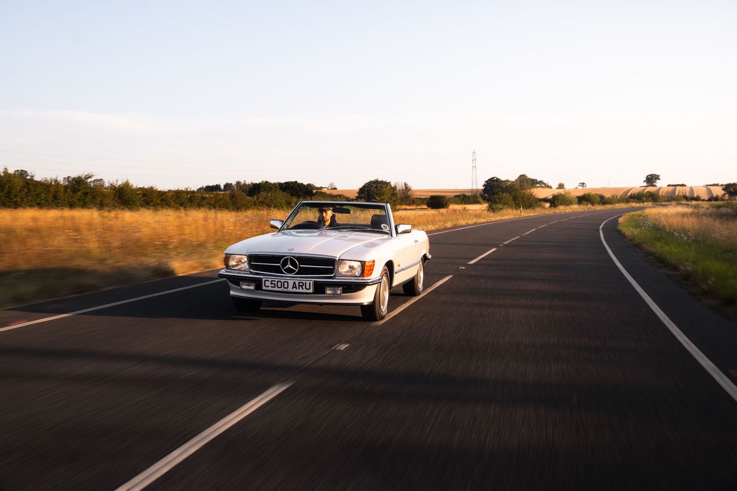 White Mercedes-Benz convertible driving on an open road at sunset, surrounded by golden fields.