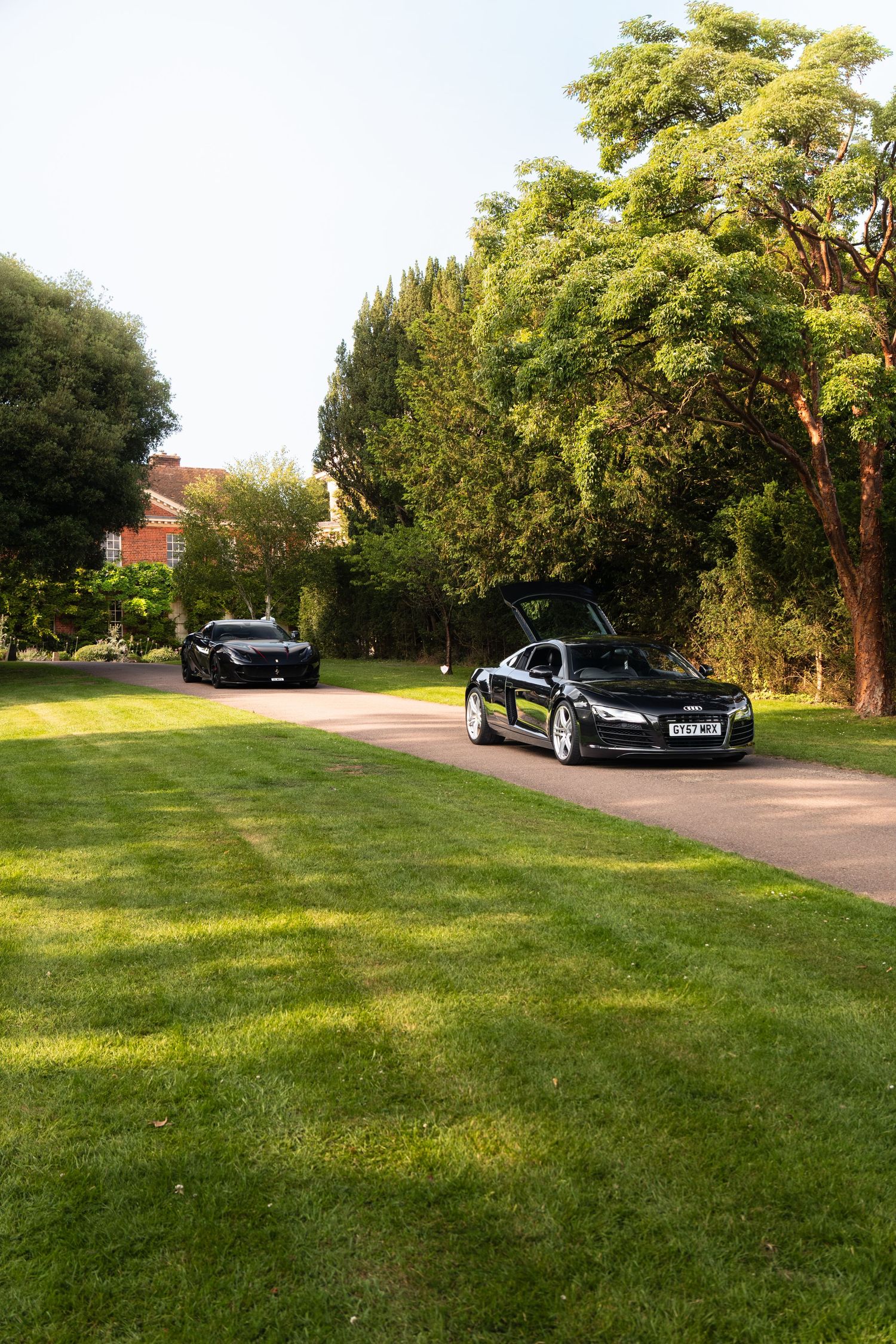 Two black luxury sports cars, including an Audi R8, parked on a driveway surrounded by lush green lawns and trees.
