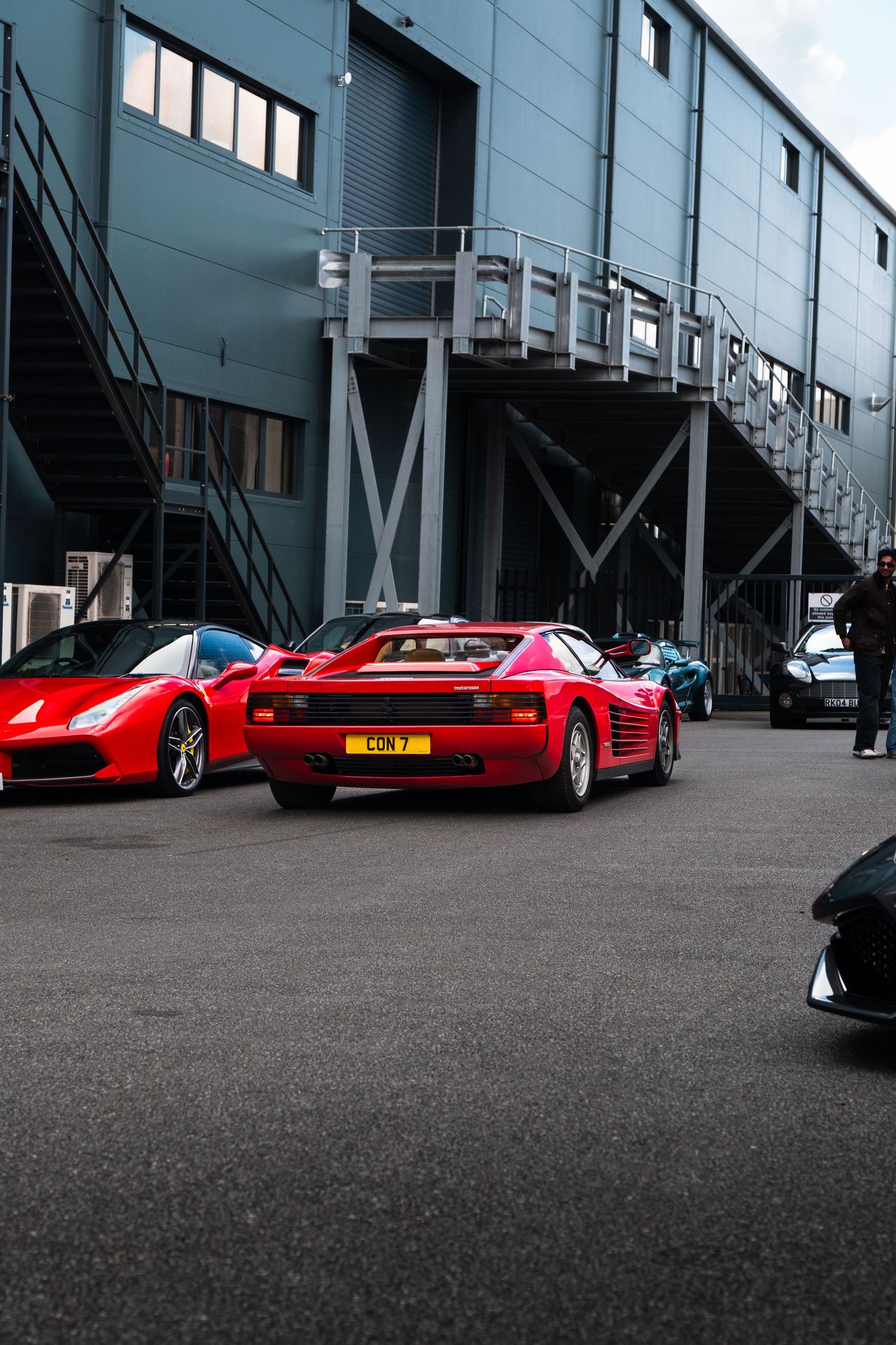 Classic red Ferrari Testarossa parked in front of an industrial building alongside other exotic supercars.