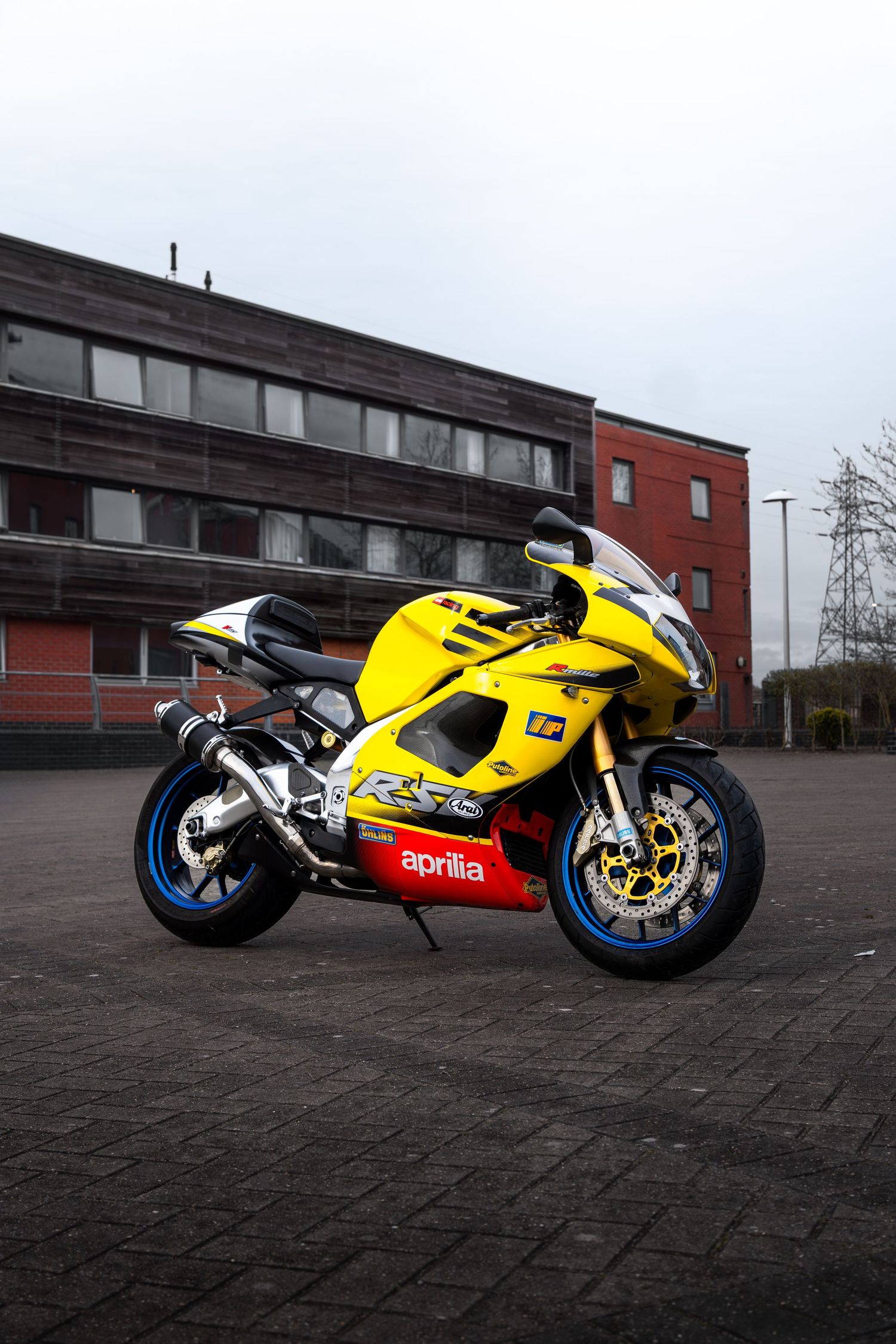 Yellow and red Aprilia RSV superbike parked on cobblestones in front of a modern brick building.