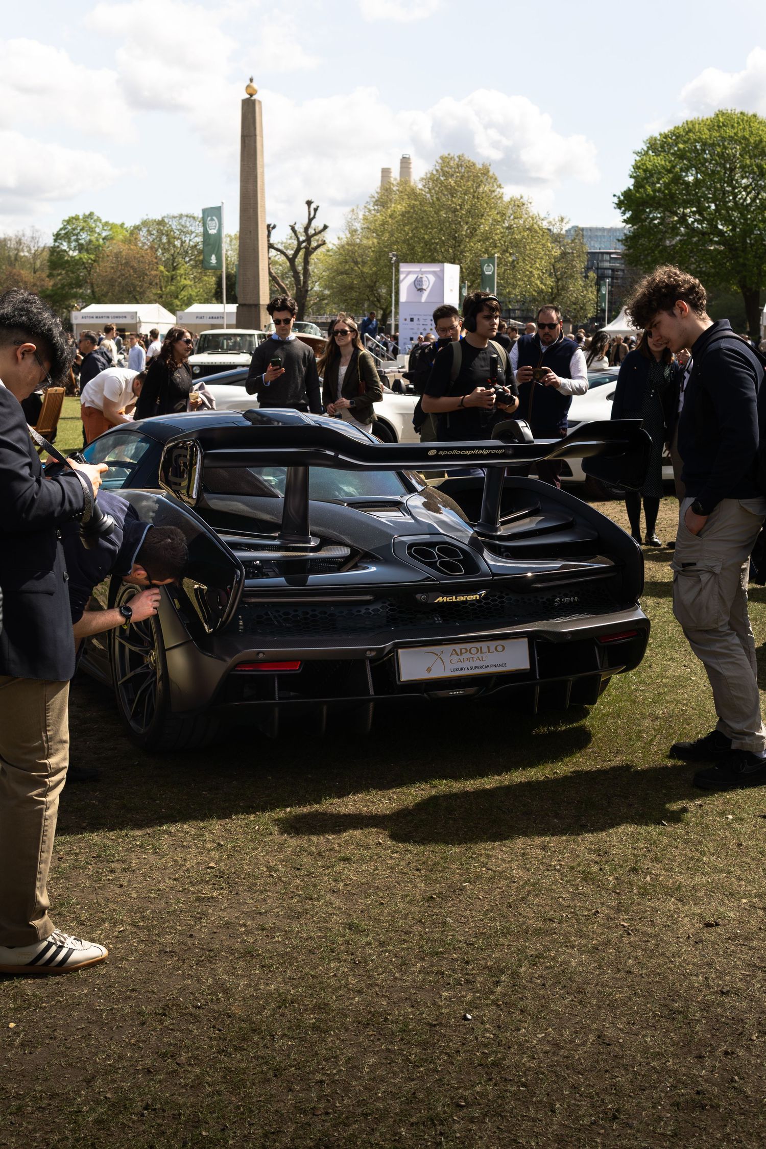 Dark McLaren supercar with large rear wing surrounded by enthusiasts at an outdoor luxury automotive event.