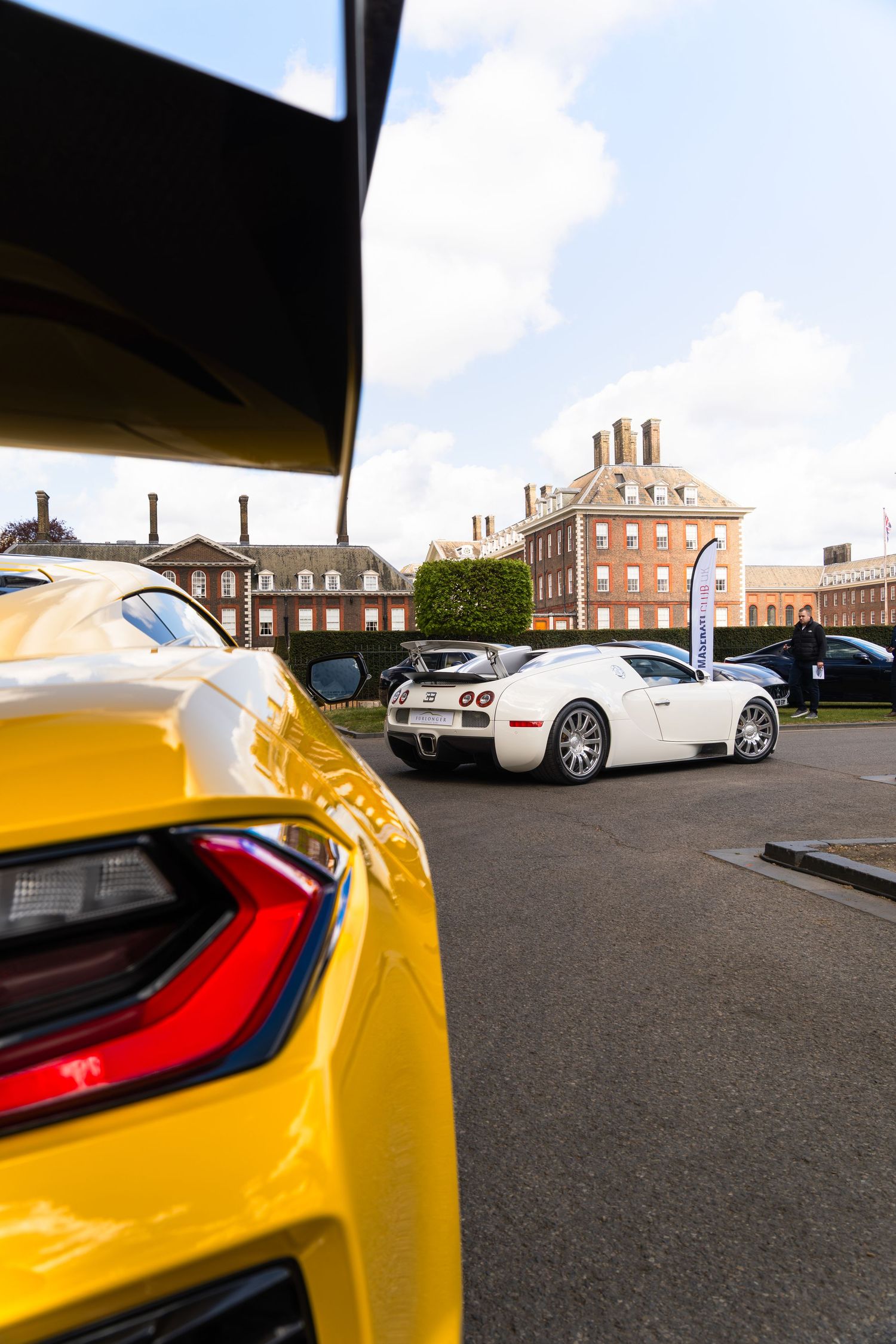 Yellow supercar and white Bugatti Veyron parked outside a grand historic brick building at a car show.