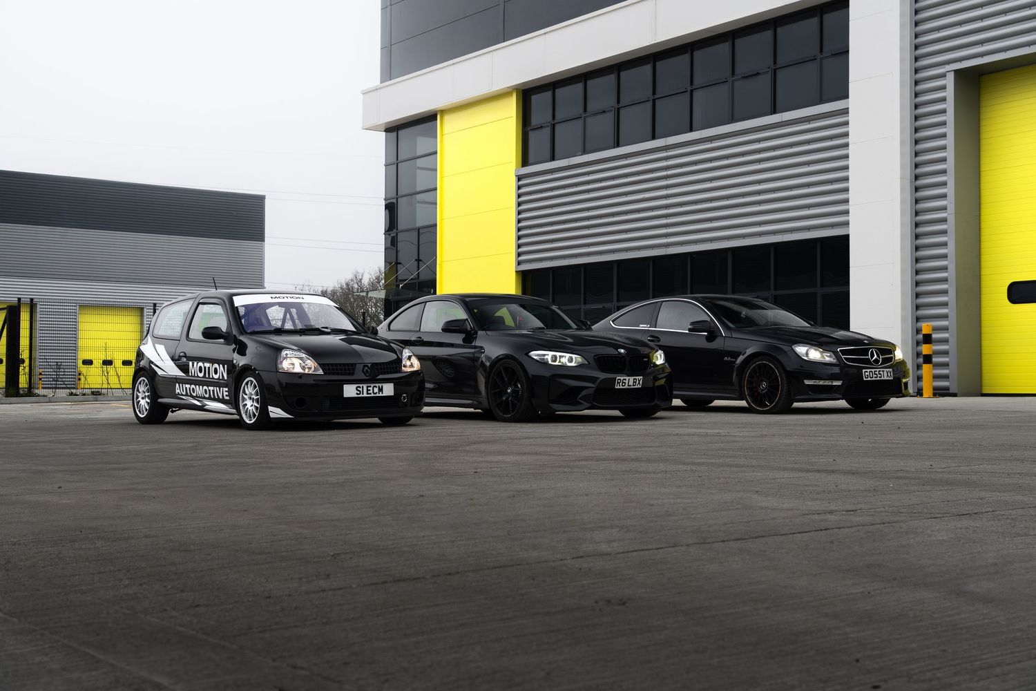 Three black Motion Automotive performance cars parked outside an industrial building with yellow doors.