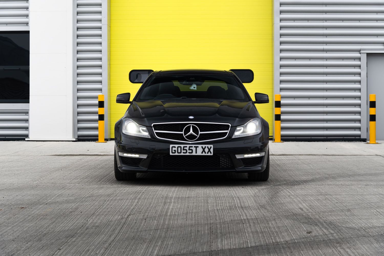 Black Mercedes-Benz C-Class coupe with GO55T XX plate parked facing forward outside yellow industrial garage doors.