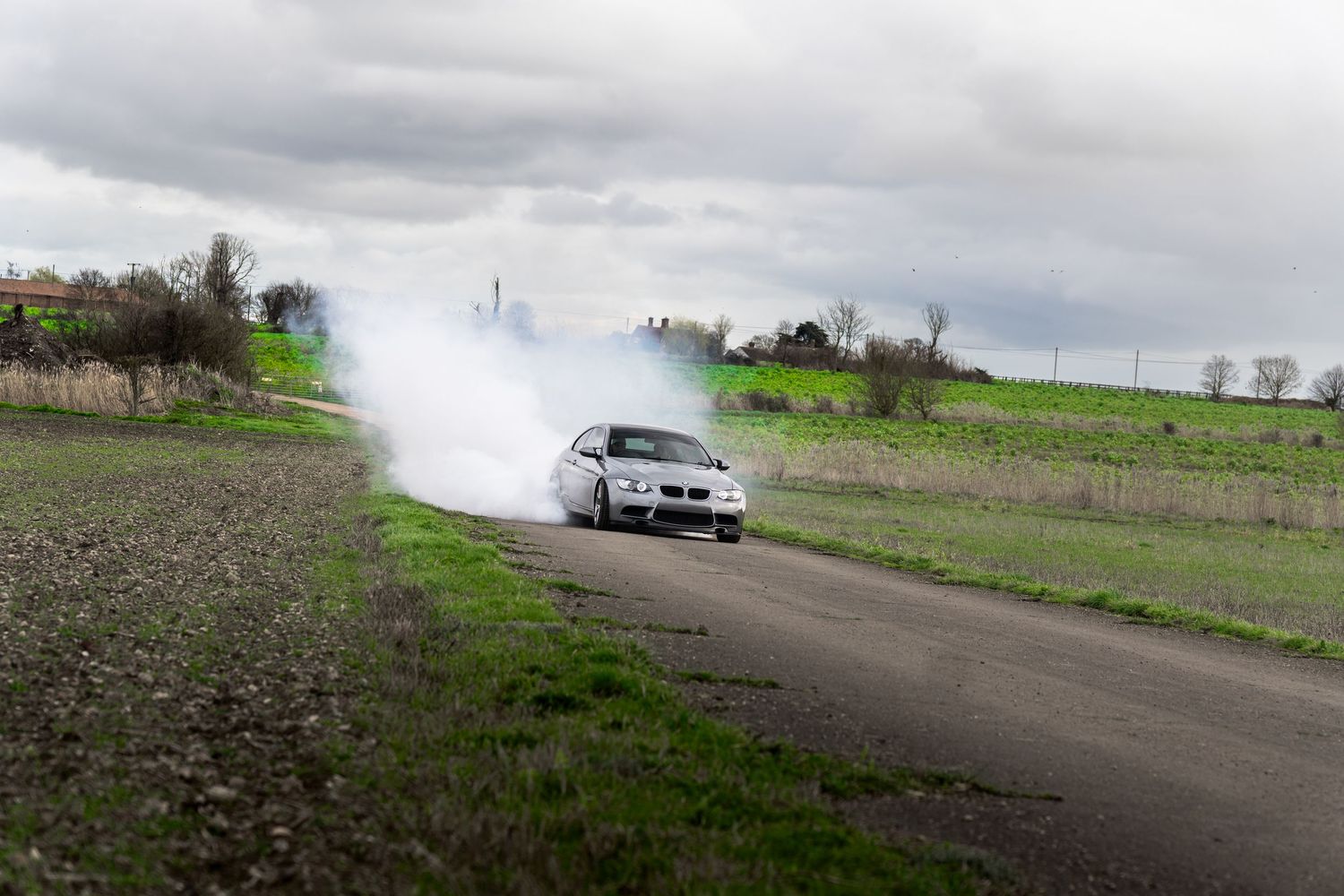 Silver BMW M3 creating large smoke clouds while drifting on a rural tarmac road.
