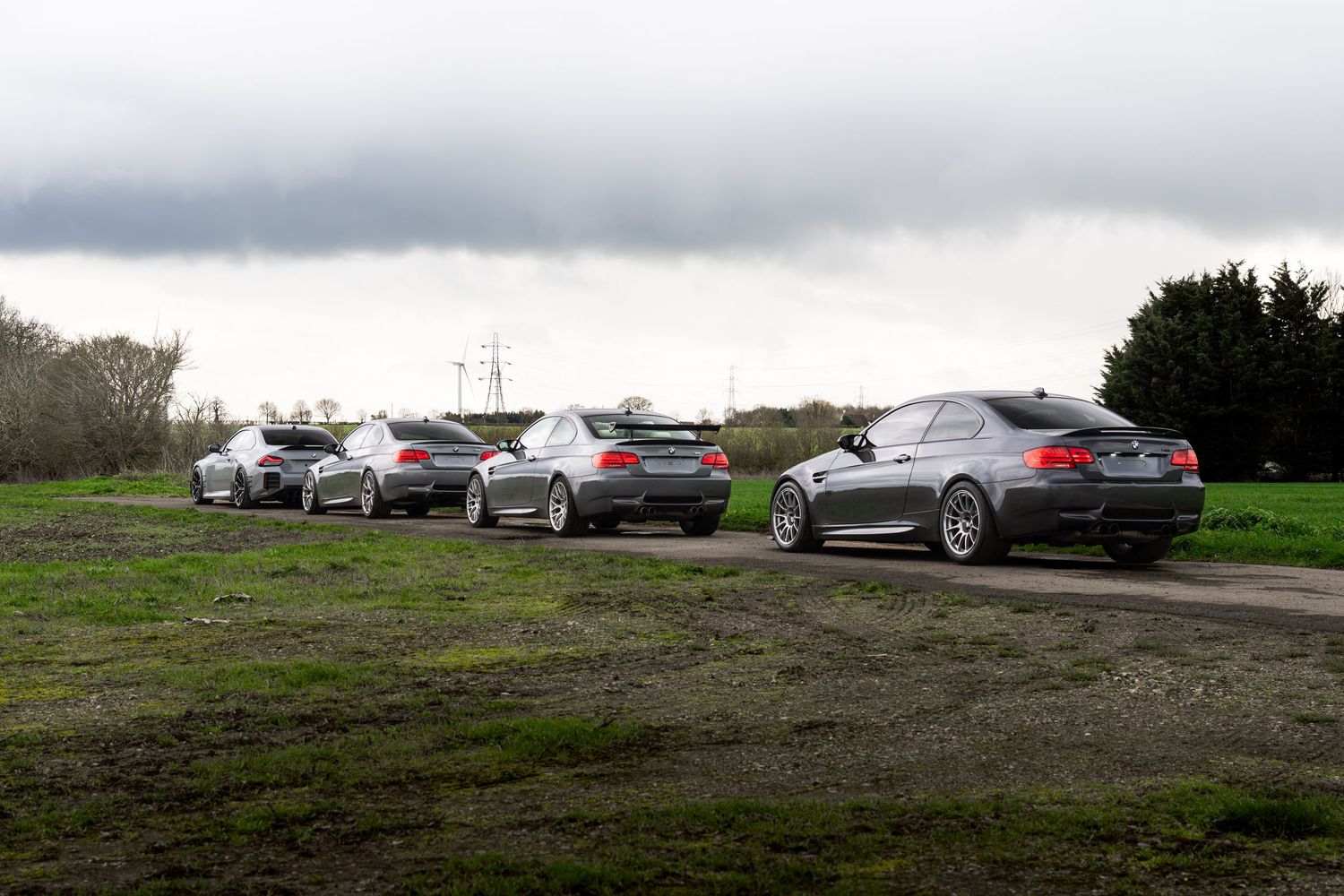 Four BMW M3 coupes parked on a dirt area with green grass under a dramatic cloudy sky.