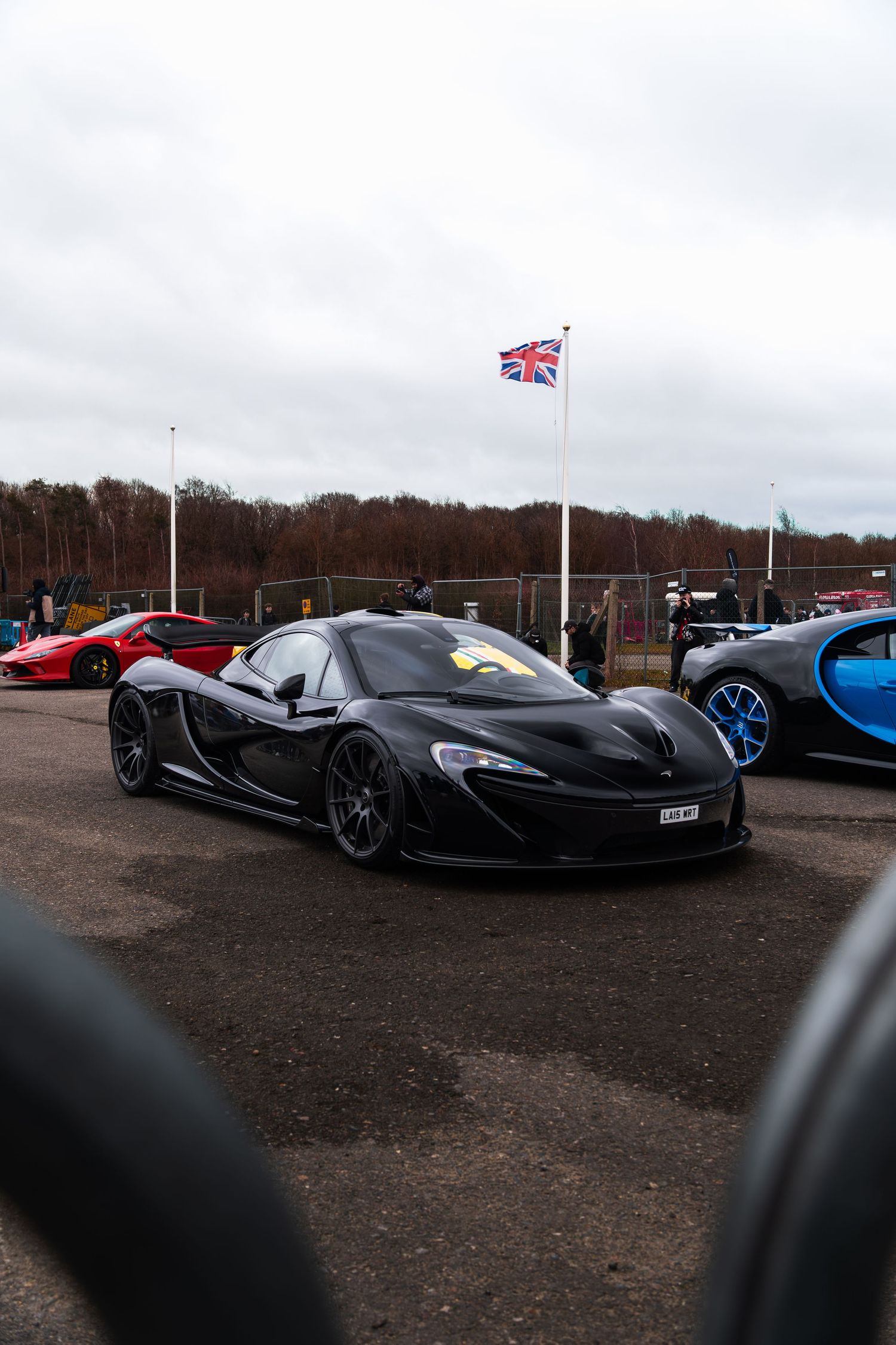 Black McLaren P1 supercar parked at a UK car meet with Union Jack flag in background.