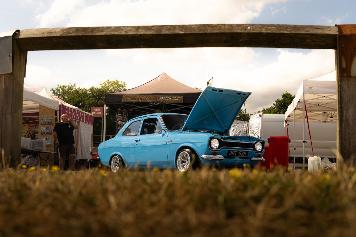 Classic blue Ford Escort MkI with open hood displayed at an outdoor car show under a wooden frame gate.