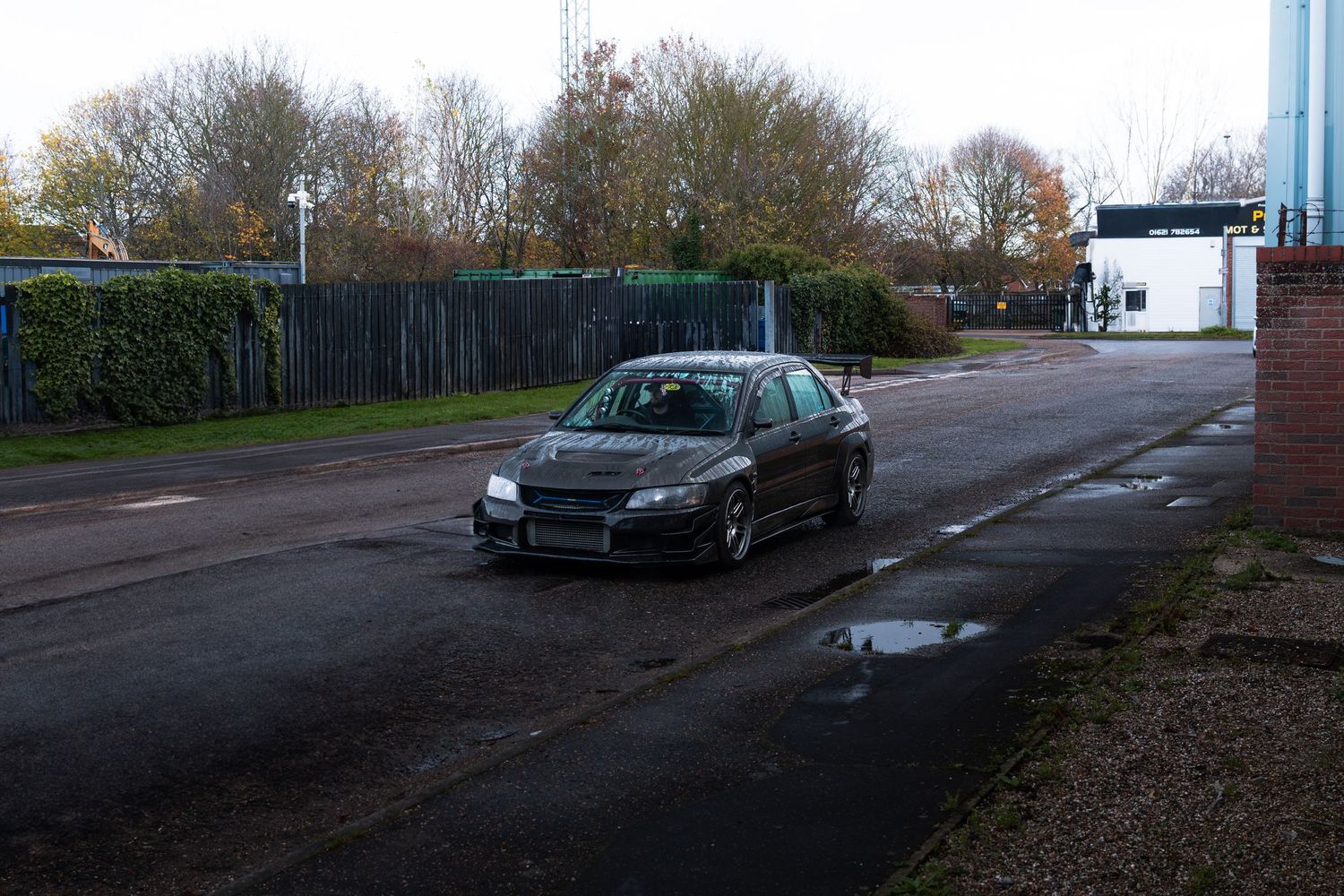 A dark Mitsubishi Lancer Evo drives along a wet road at an industrial estate in autumn.