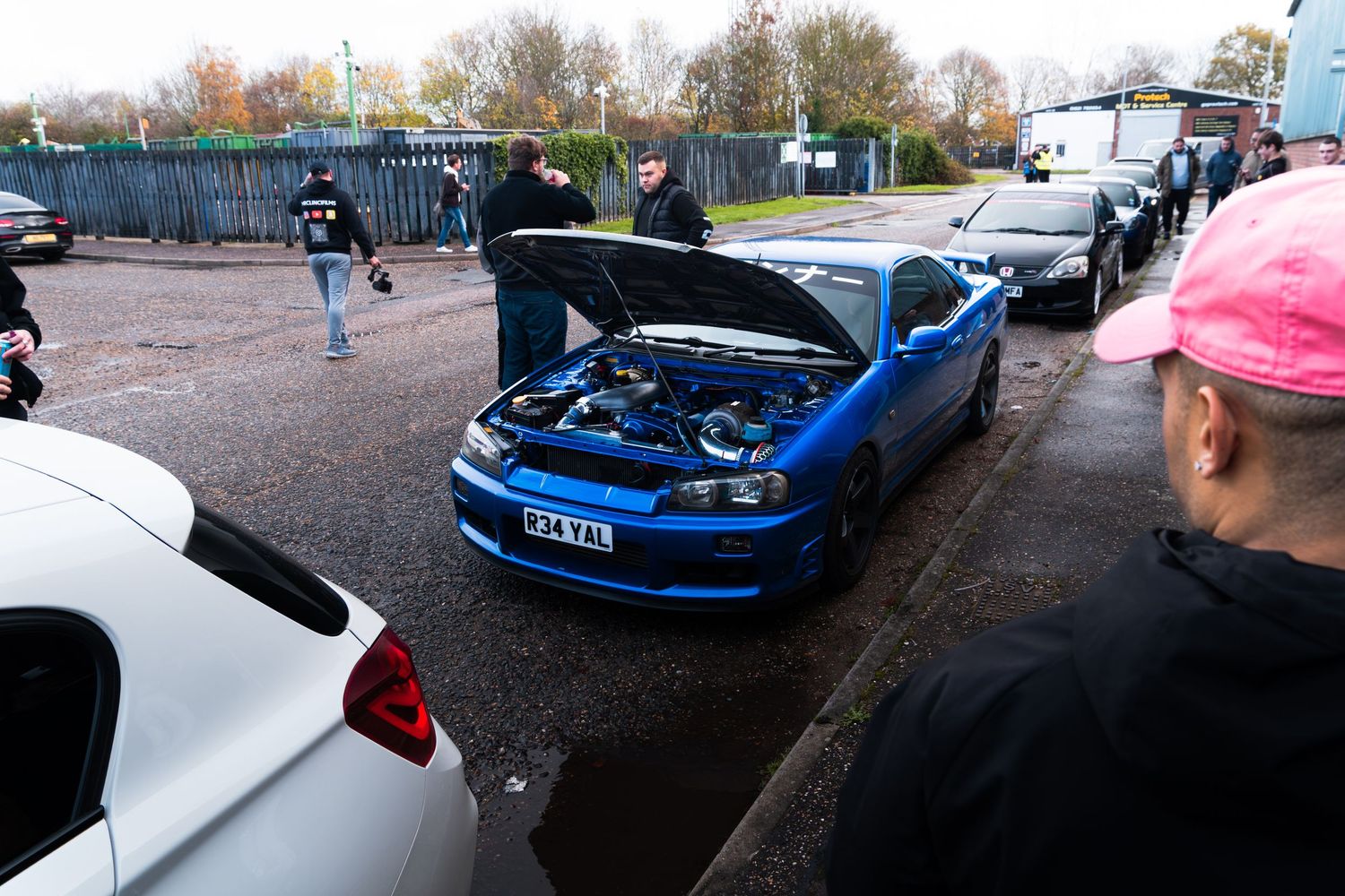 Blue modified car with hood open at outdoor car meet, surrounded by enthusiasts in a UK car park.