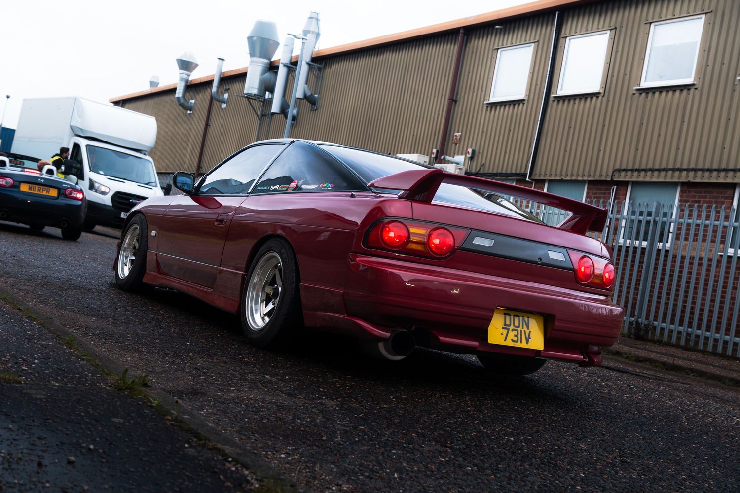Rear view of a modified red Nissan 200SX with a spoiler parked outside an industrial building.